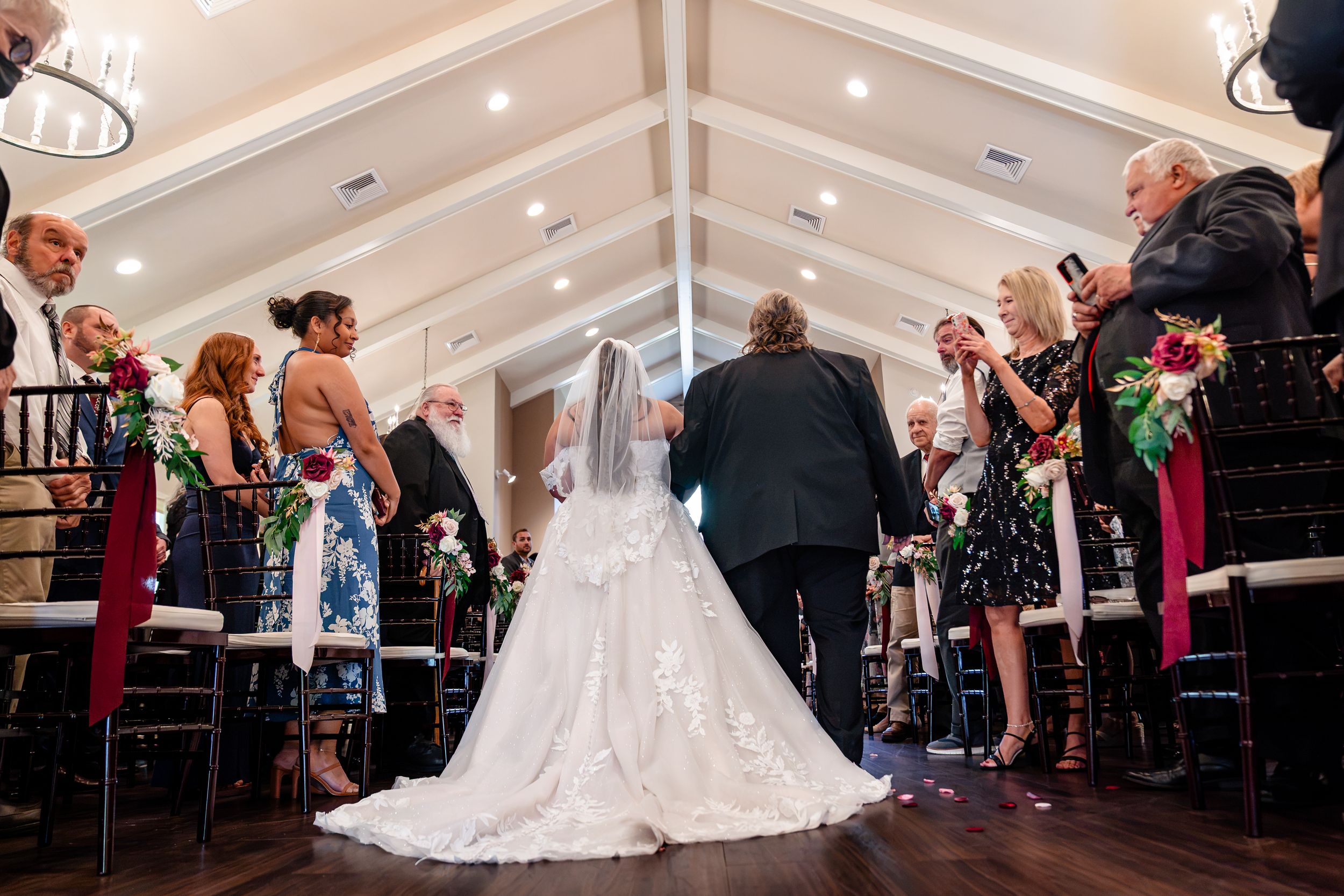 Bride in lace gown walking down aisle with escort at elegant indoor wedding ceremony.