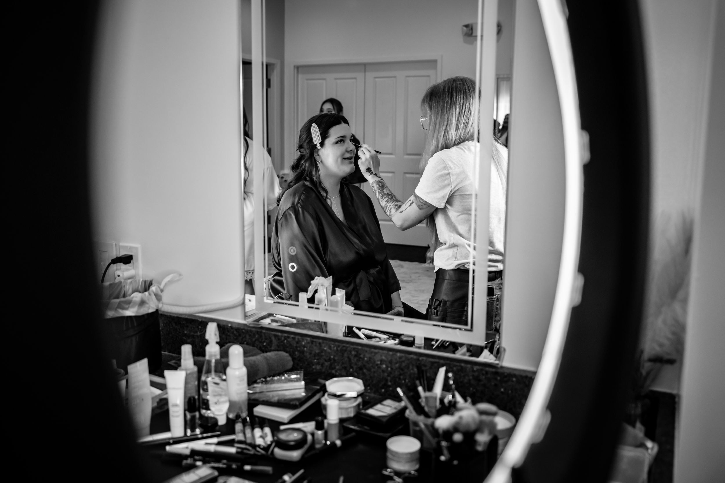 Black and white photo of a bride getting makeup applied by artist, reflected in a vanity mirror surrounded by beauty products.