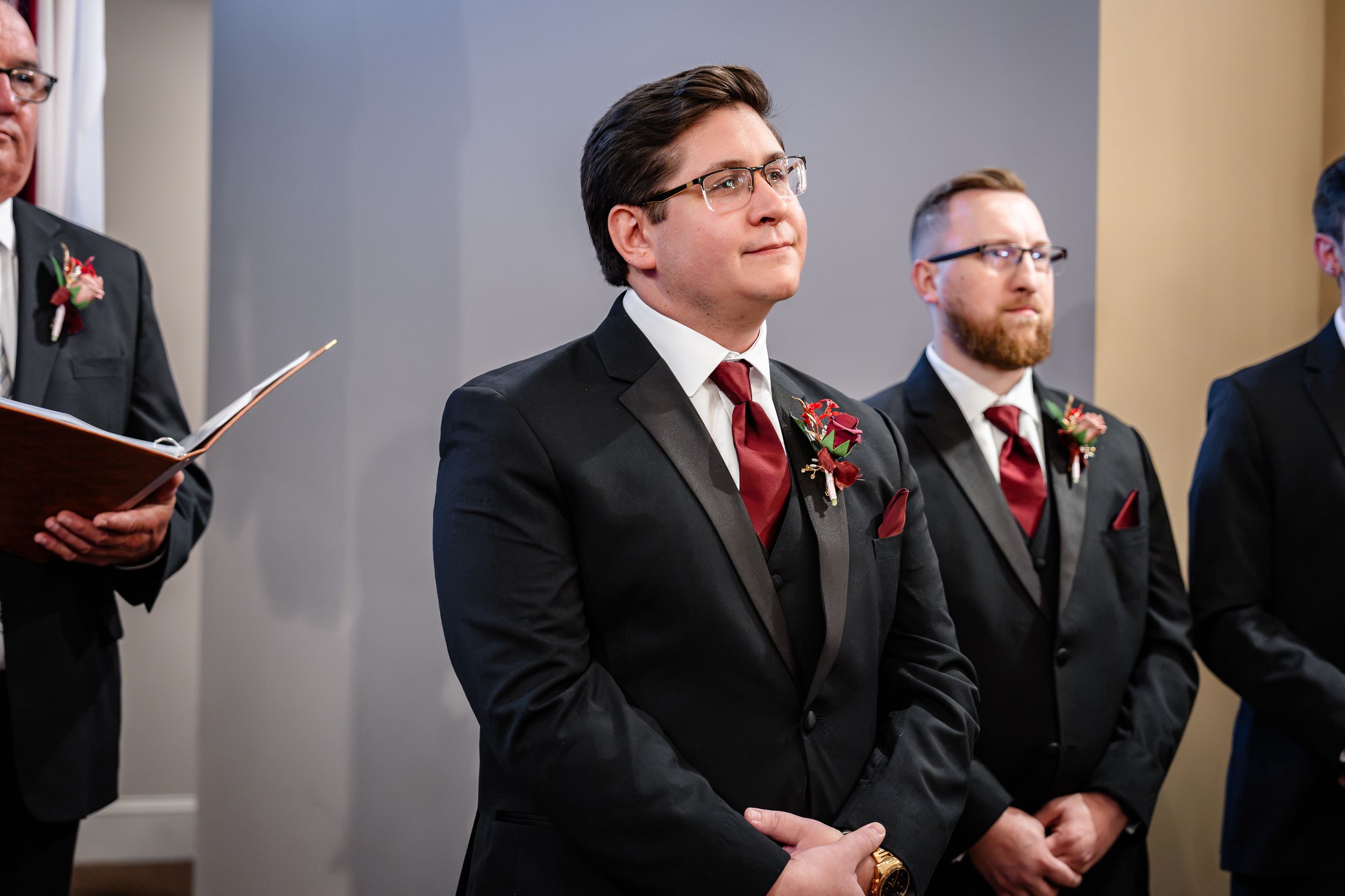 Groom in black suit with red tie stands at altar during ceremony.