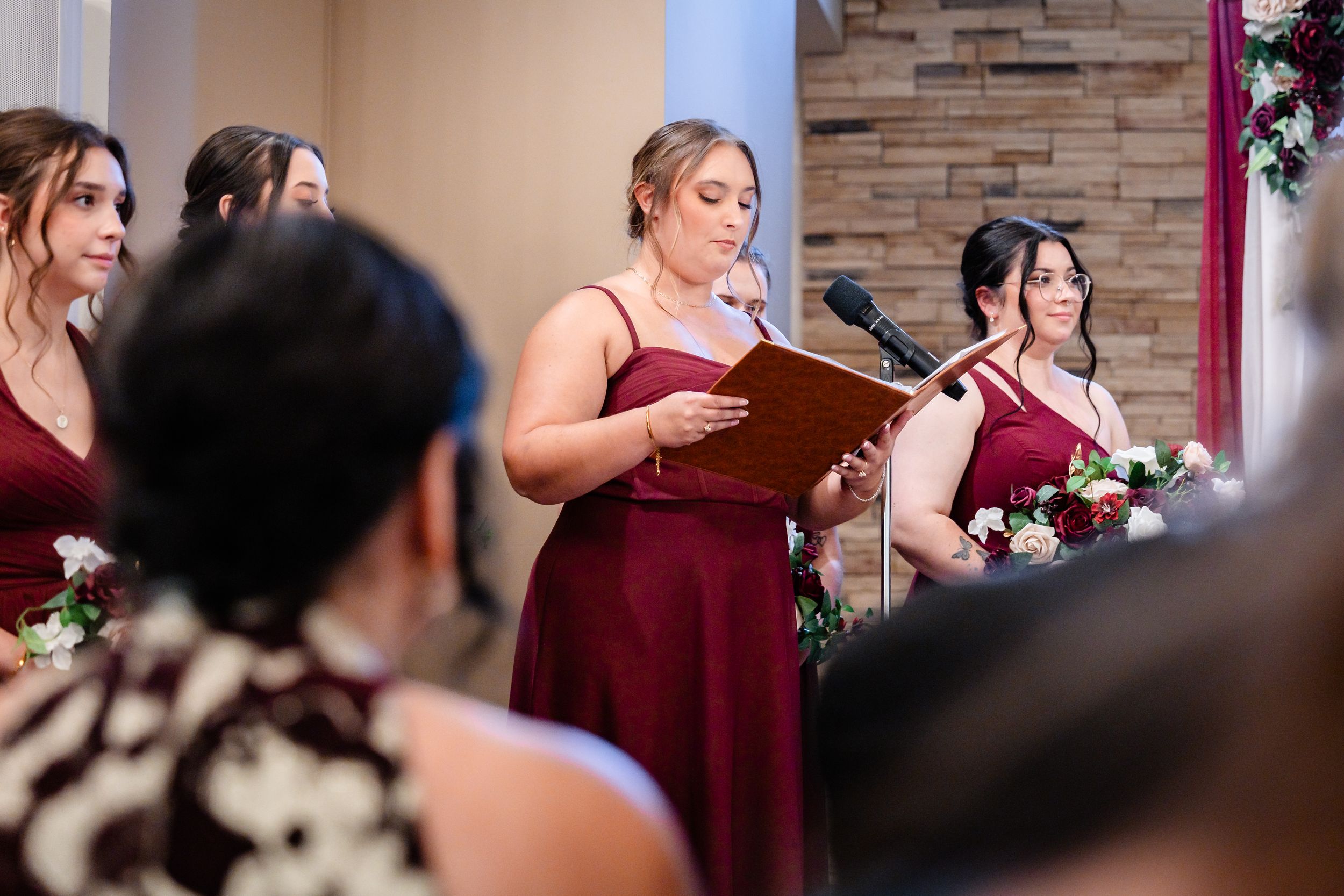 Bridesmaids in burgundy dresses stand at wedding altar, one reading from a book during the ceremony.