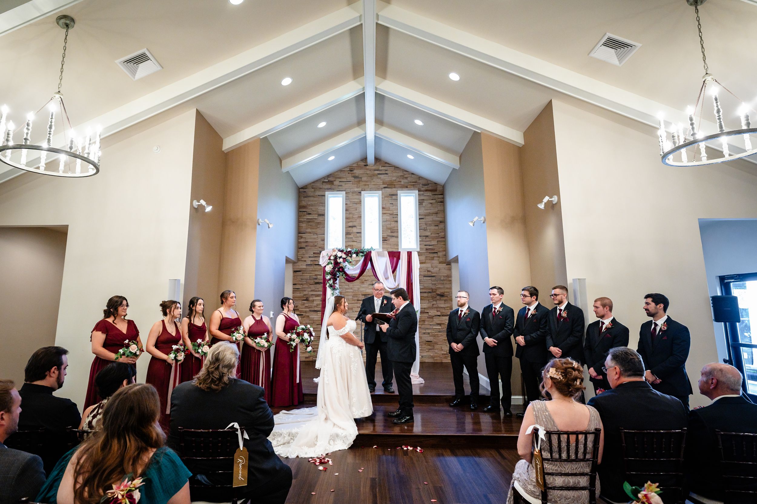 Wedding ceremony in chapel with bridesmaids in burgundy dresses and groomsmen at the altar.