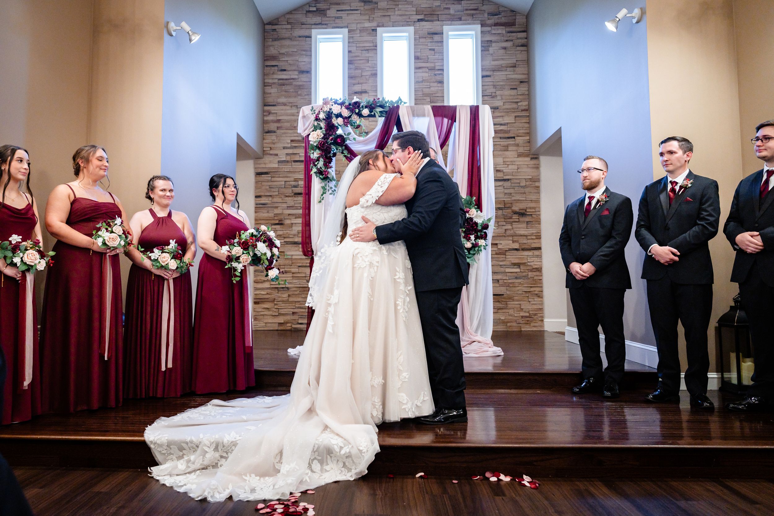 Bride and groom share first kiss at wedding altar surrounded by bridesmaids in burgundy dresses and groomsmen.