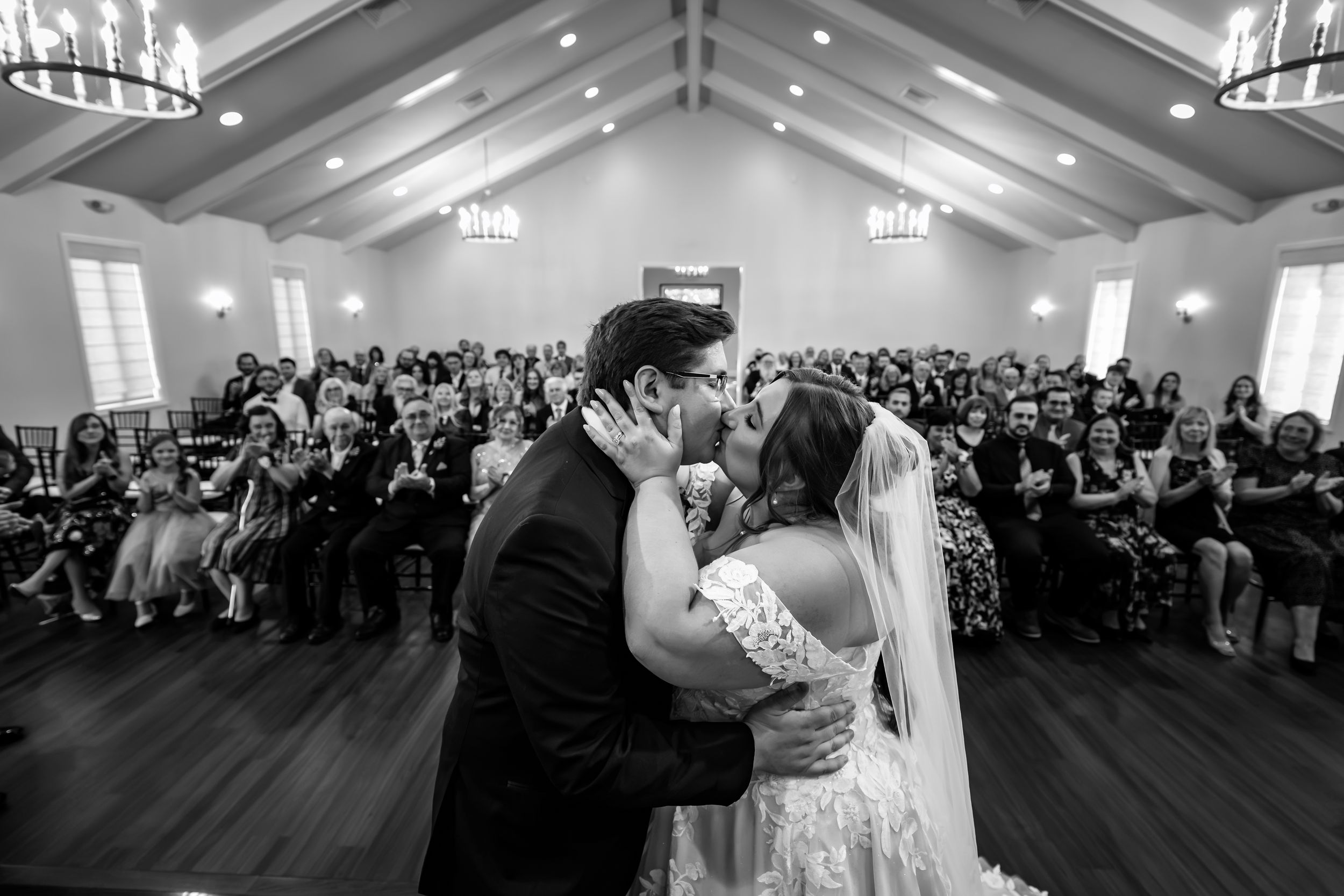 Black and white photo of newlyweds sharing first kiss before guests.