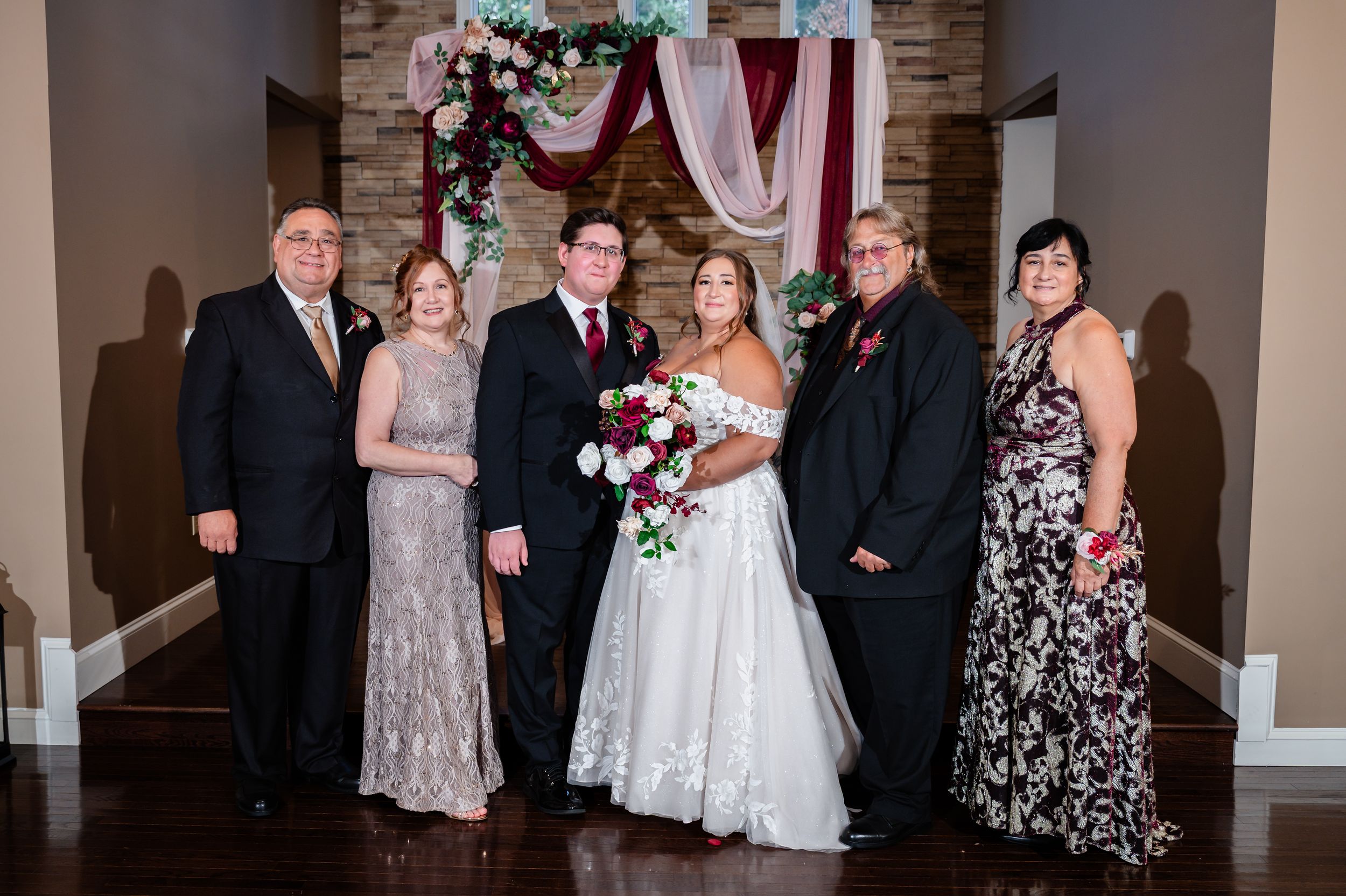 Wedding couple posing with four family members under floral arch in elegant indoor venue.