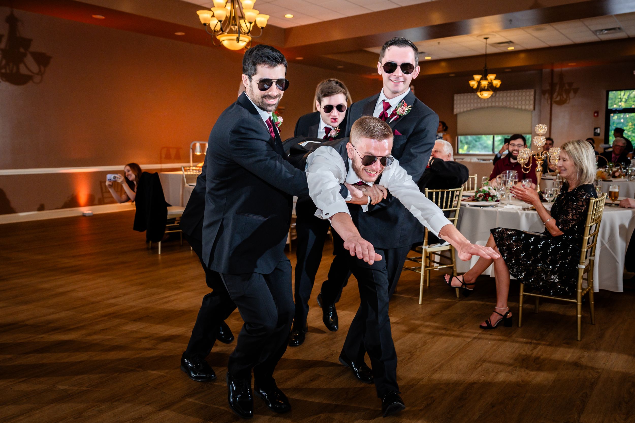 Four men in suits wearing sunglasses striking fun poses on wedding reception dance floor.