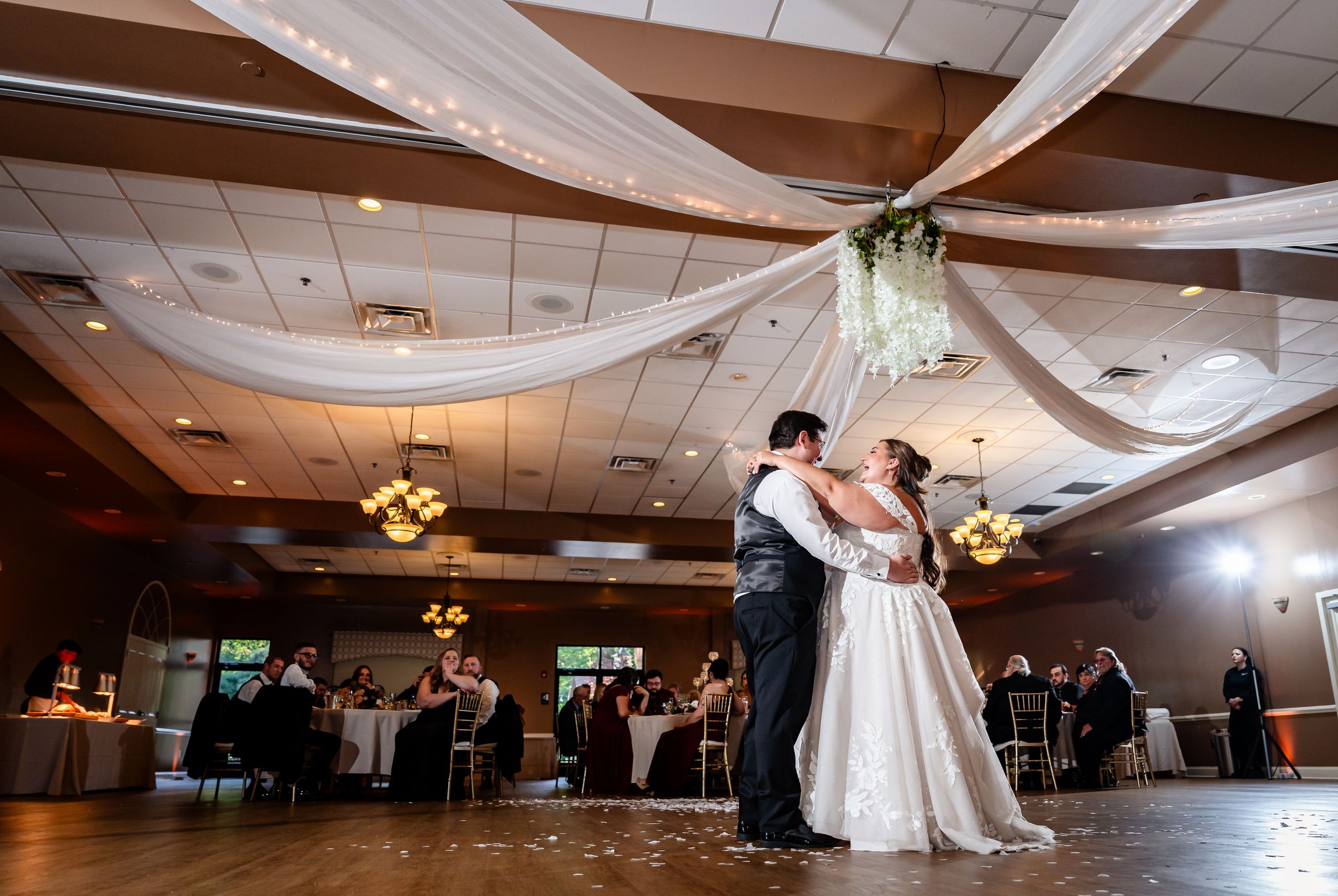 Bride and groom share their first dance under draped white fabric ceiling in a warmly lit wedding reception hall.
