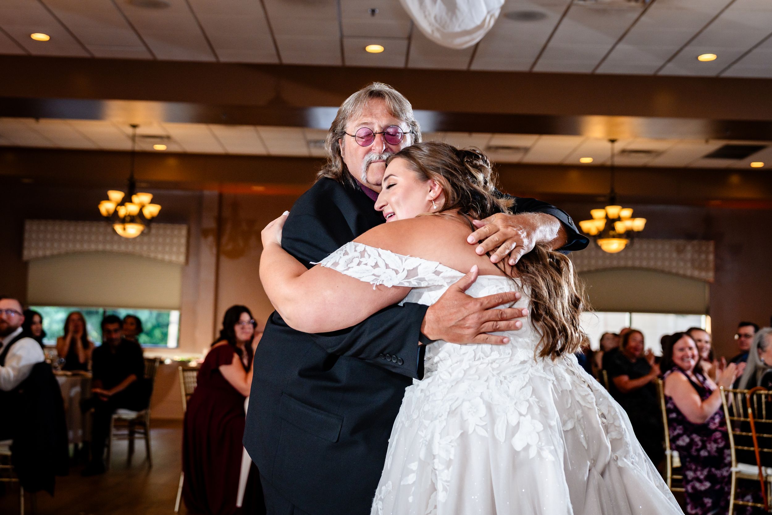 Bride in lace gown sharing warm embrace with older woman on wedding reception dance floor.