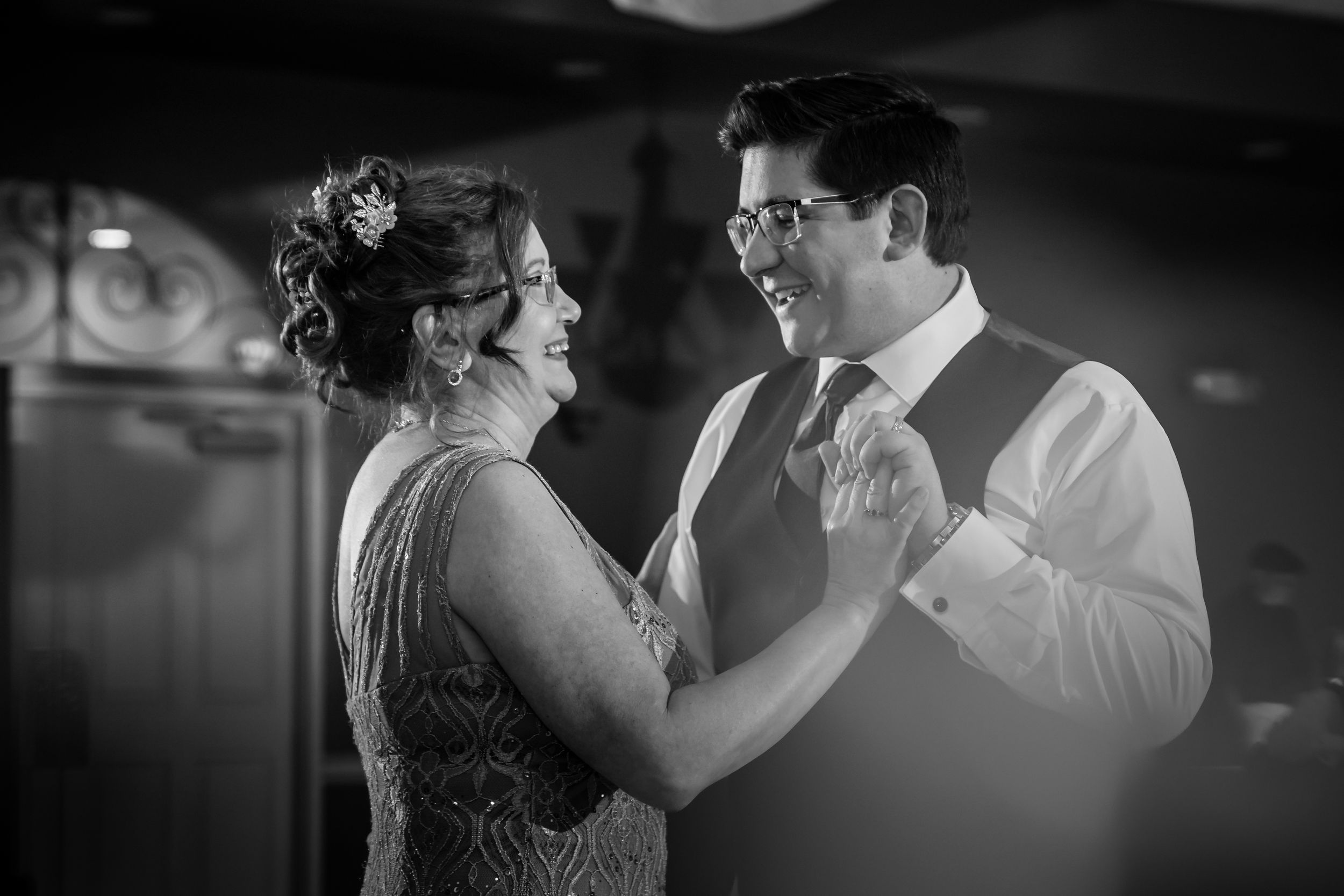 Black and white photo of groom sharing first dance with mother in elegant beaded dress at wedding reception.
