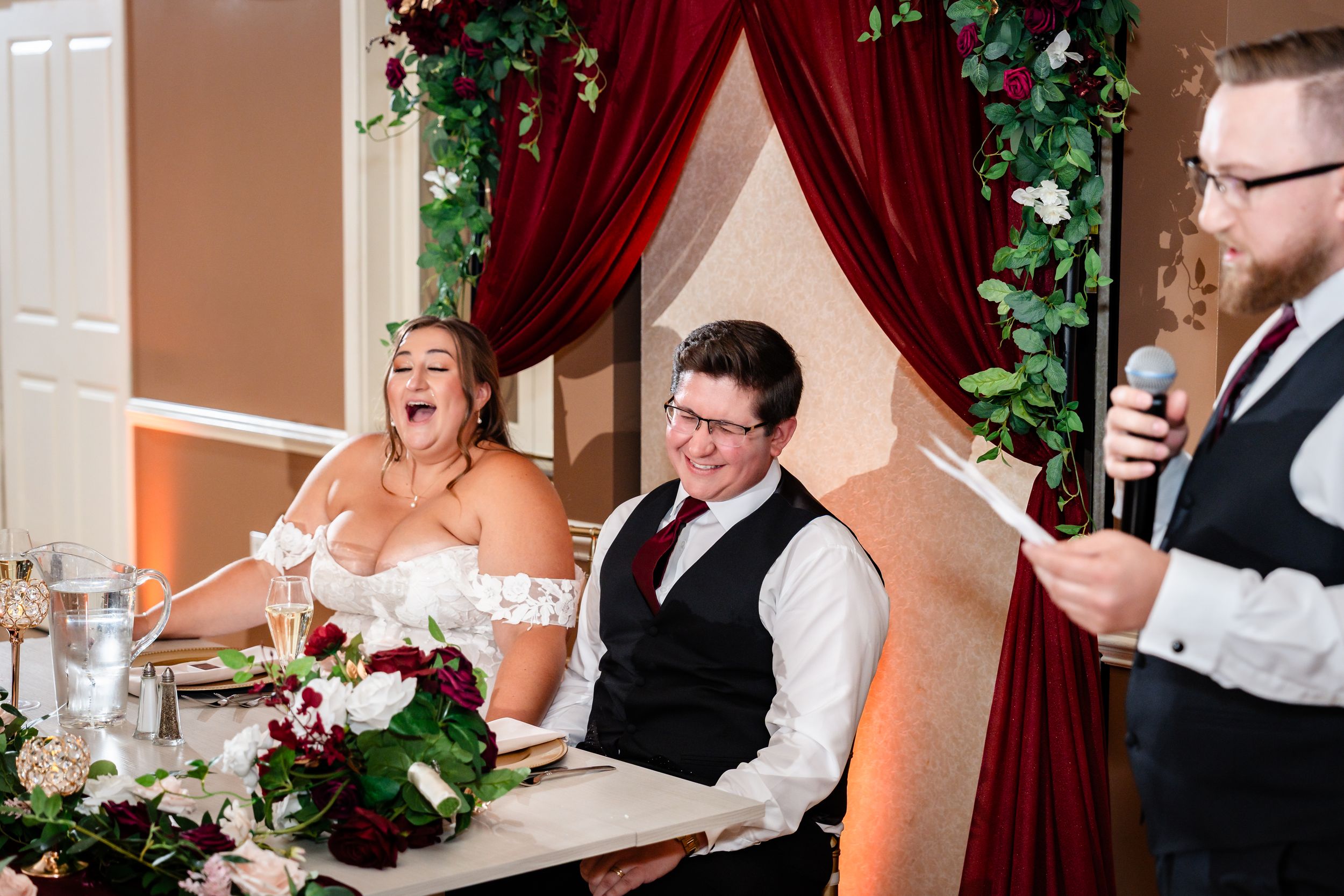 Bride laughing joyfully beside groom as a groomsman delivers a speech at the wedding reception sweetheart table.