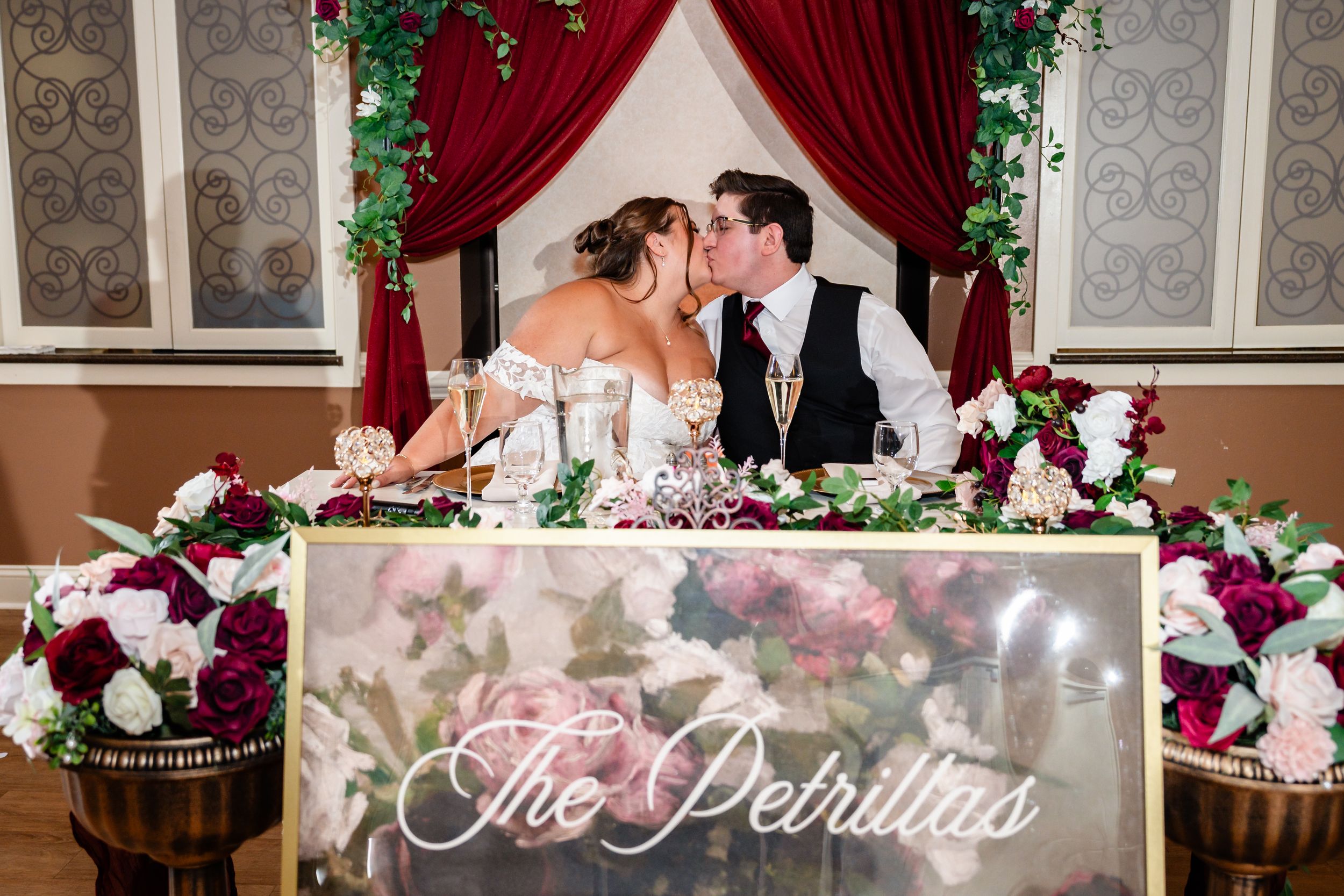 Newlyweds share a kiss at their sweetheart table adorned with burgundy and white floral arrangements at The Petrillas wedding.