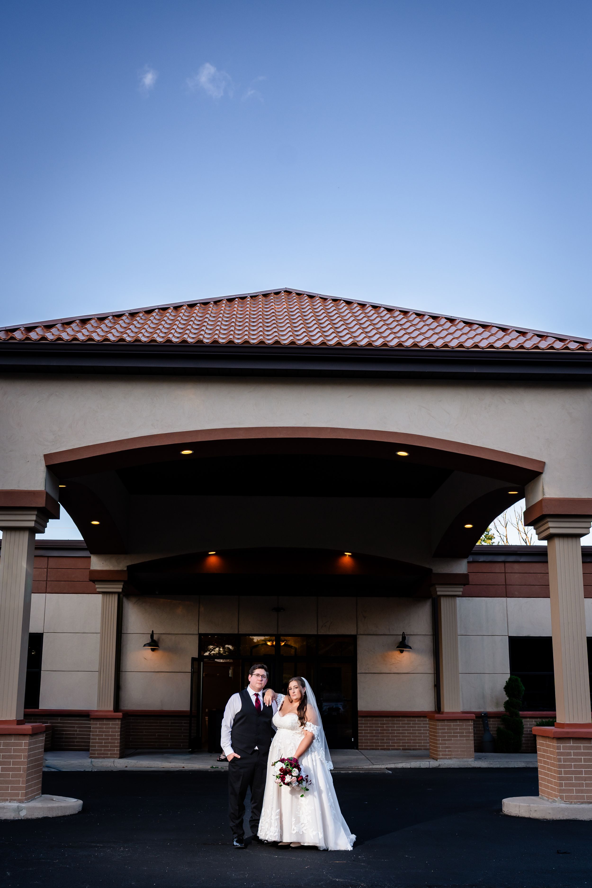 Bride and groom pose together in front of an elegant venue entrance under a blue evening sky.