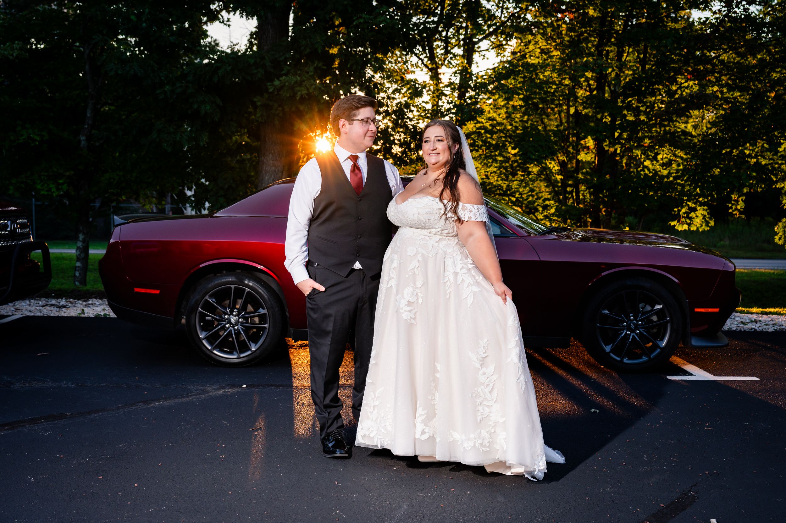 Bride and groom posing together in front of a dark red Dodge Challenger at sunset.