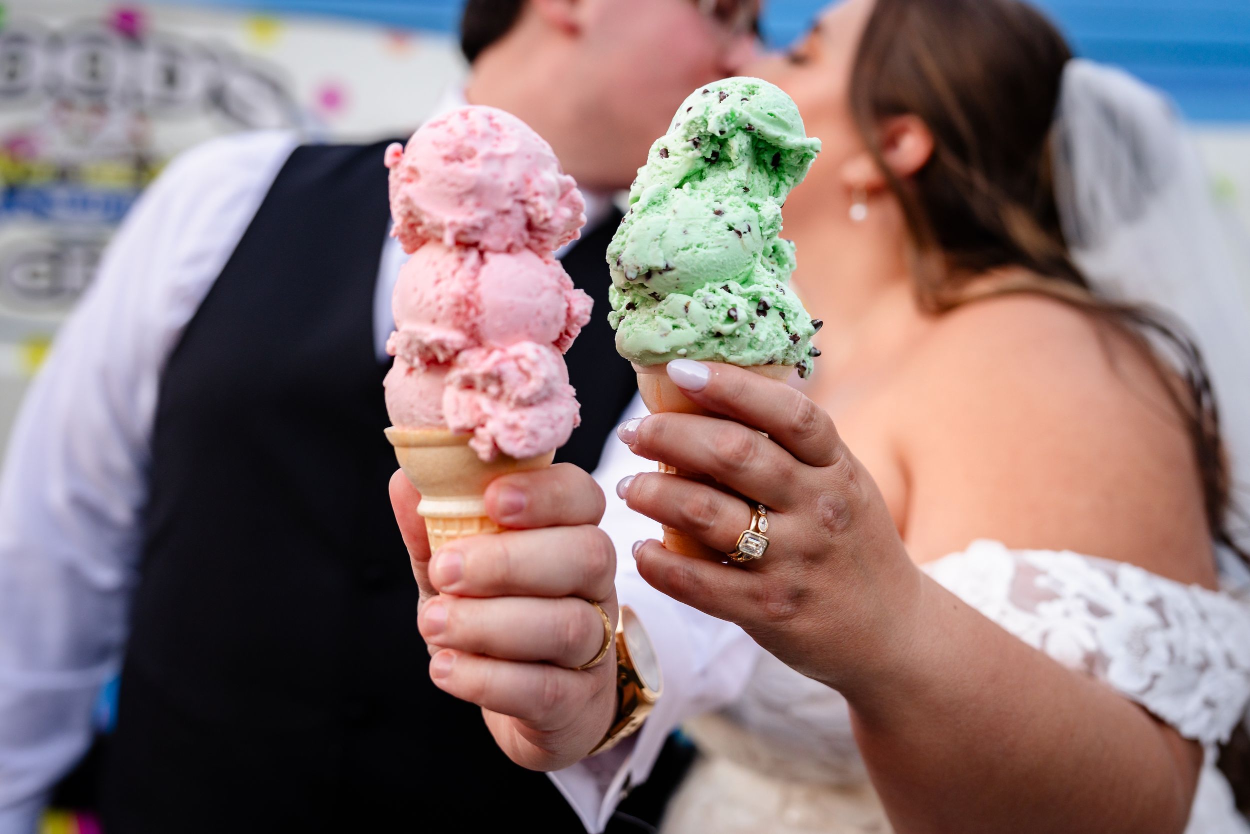 Bride and groom share a kiss while holding large pink and mint green ice cream cones at their wedding celebration.