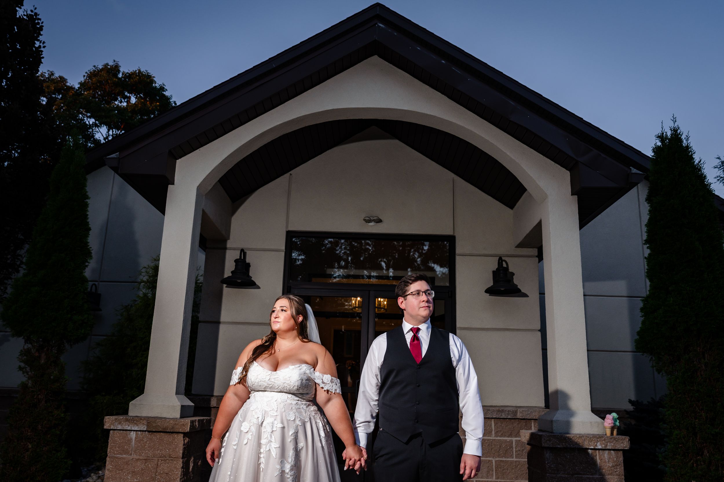 Bride and groom hold hands standing before a white chapel entrance at dusk during their wedding celebration.