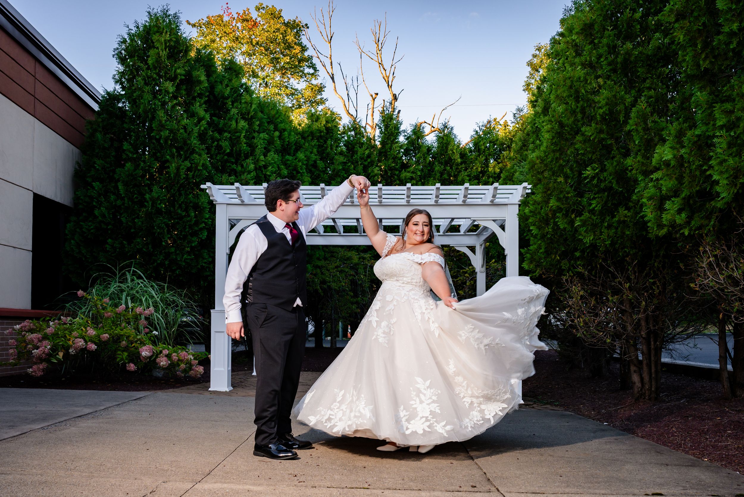 Bride and groom dancing outdoors near a white pergola surrounded by lush green trees.