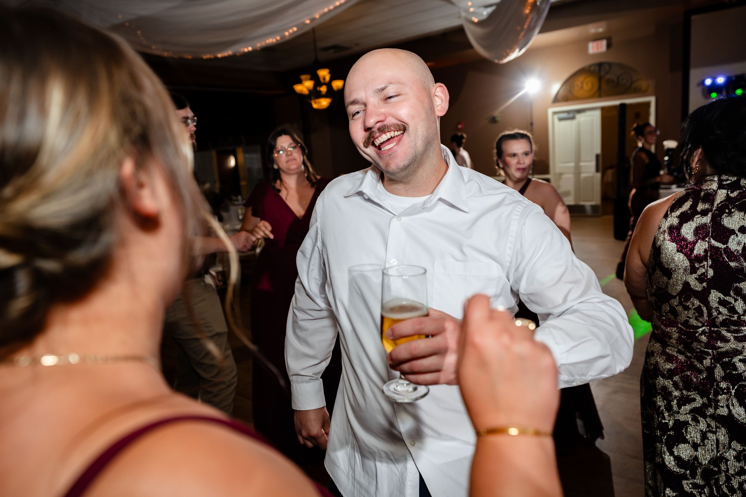 Bald man with mustache laughs and dances holding a beer at a lively wedding reception.