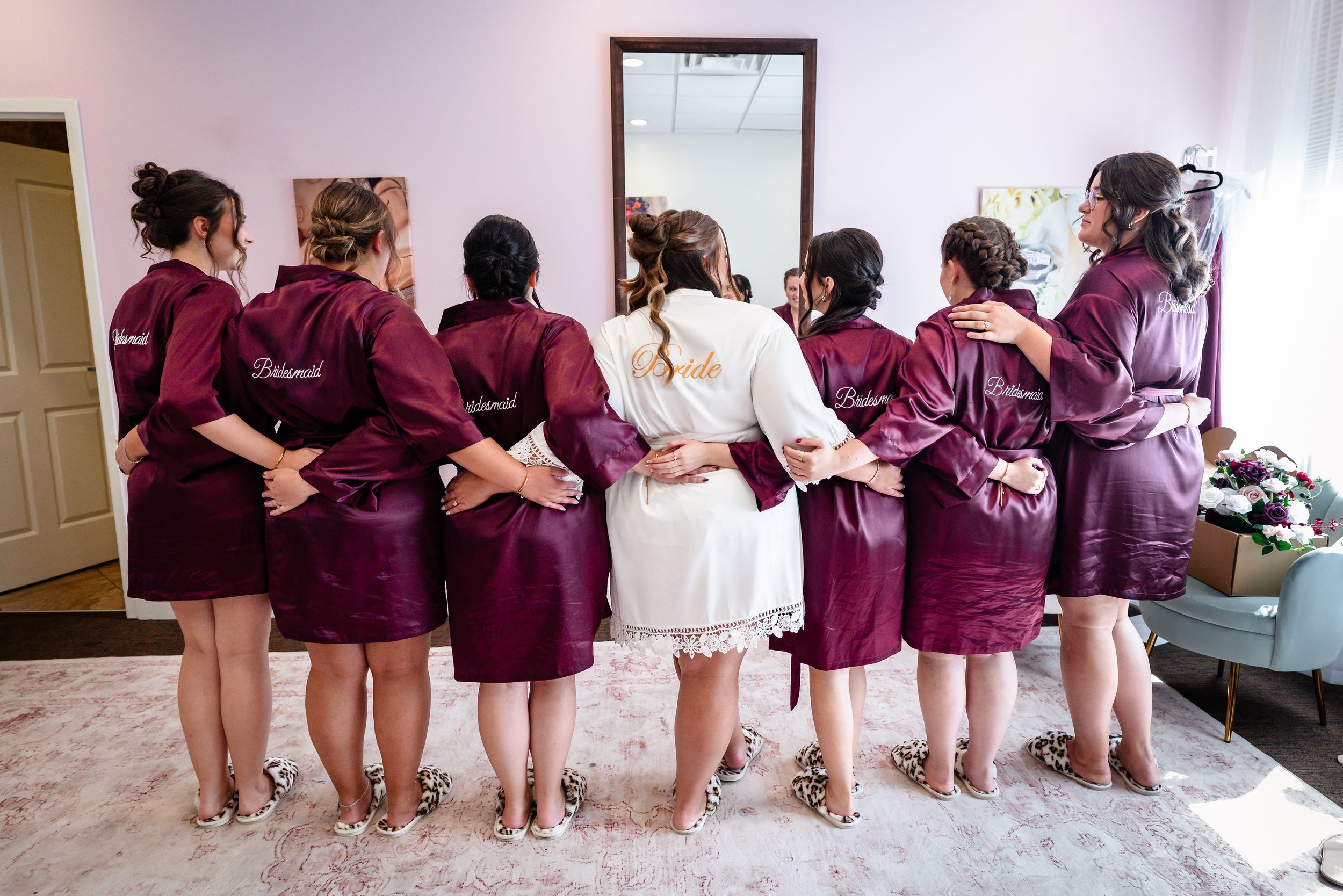 Bride and bridesmaids in matching burgundy satin robes standing together facing a mirror during wedding preparations.