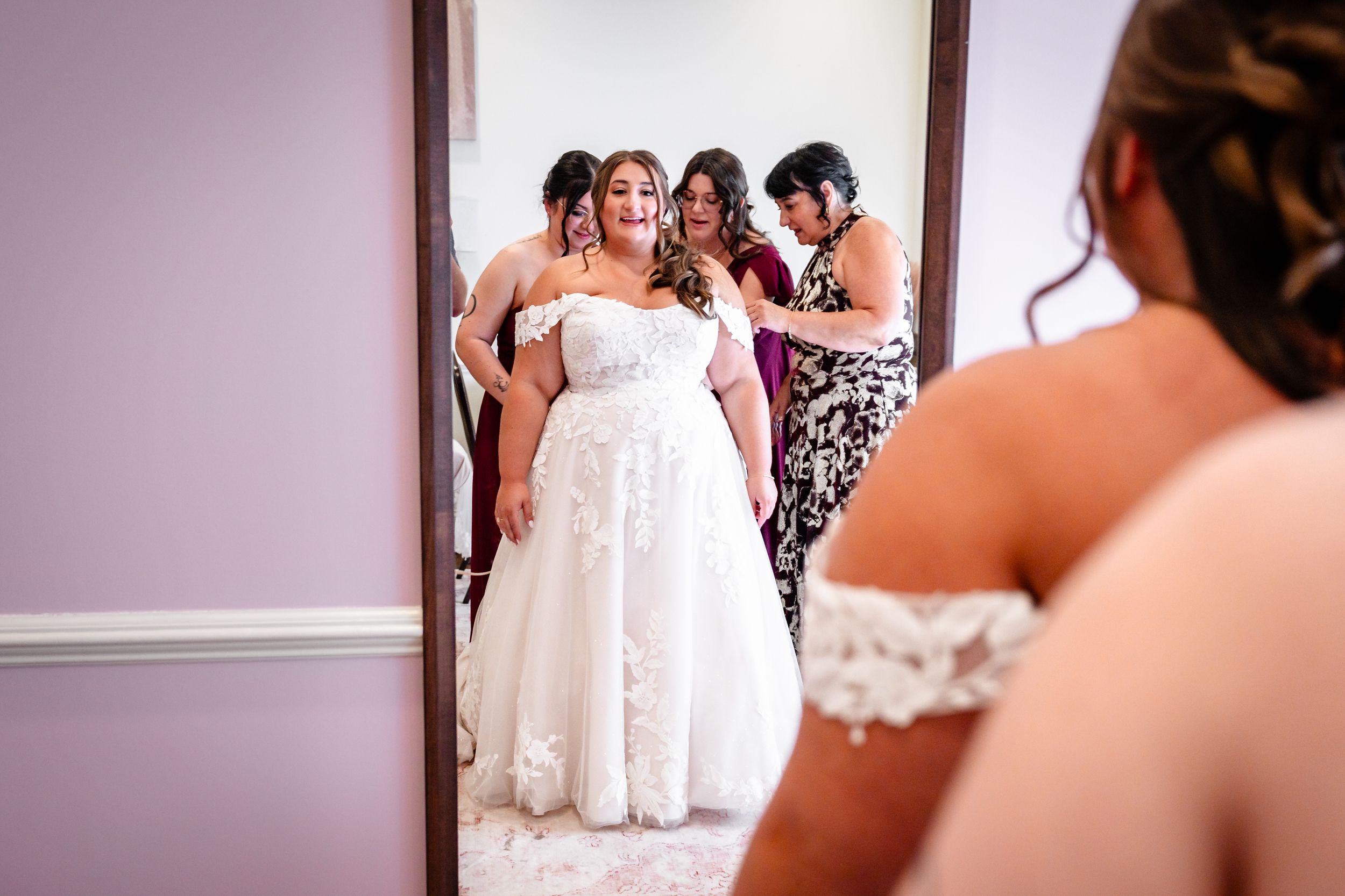Bride in lace wedding gown being helped by bridesmaids while viewing her reflection in a full-length mirror.