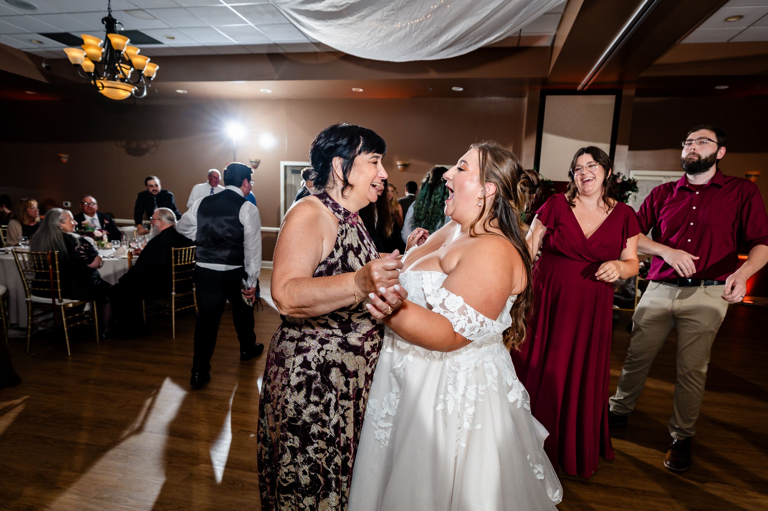Bride in white gown dancing and laughing with a guest in floral dress at a lively wedding reception.