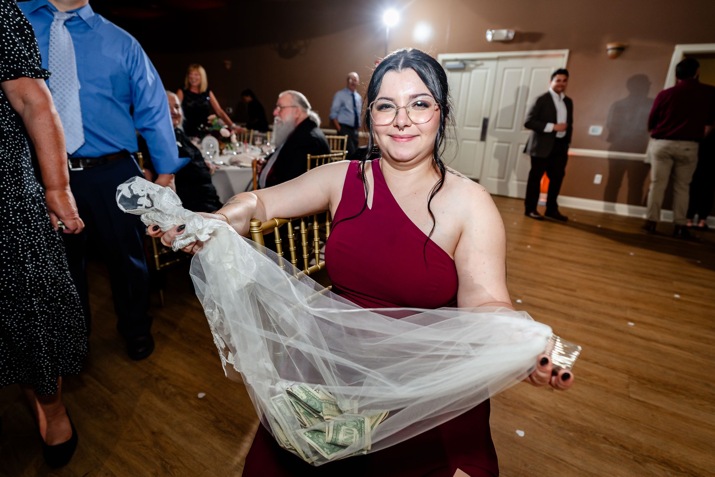 Bridesmaid in burgundy dress smiling while holding the bride's veil on the wedding reception dance floor.