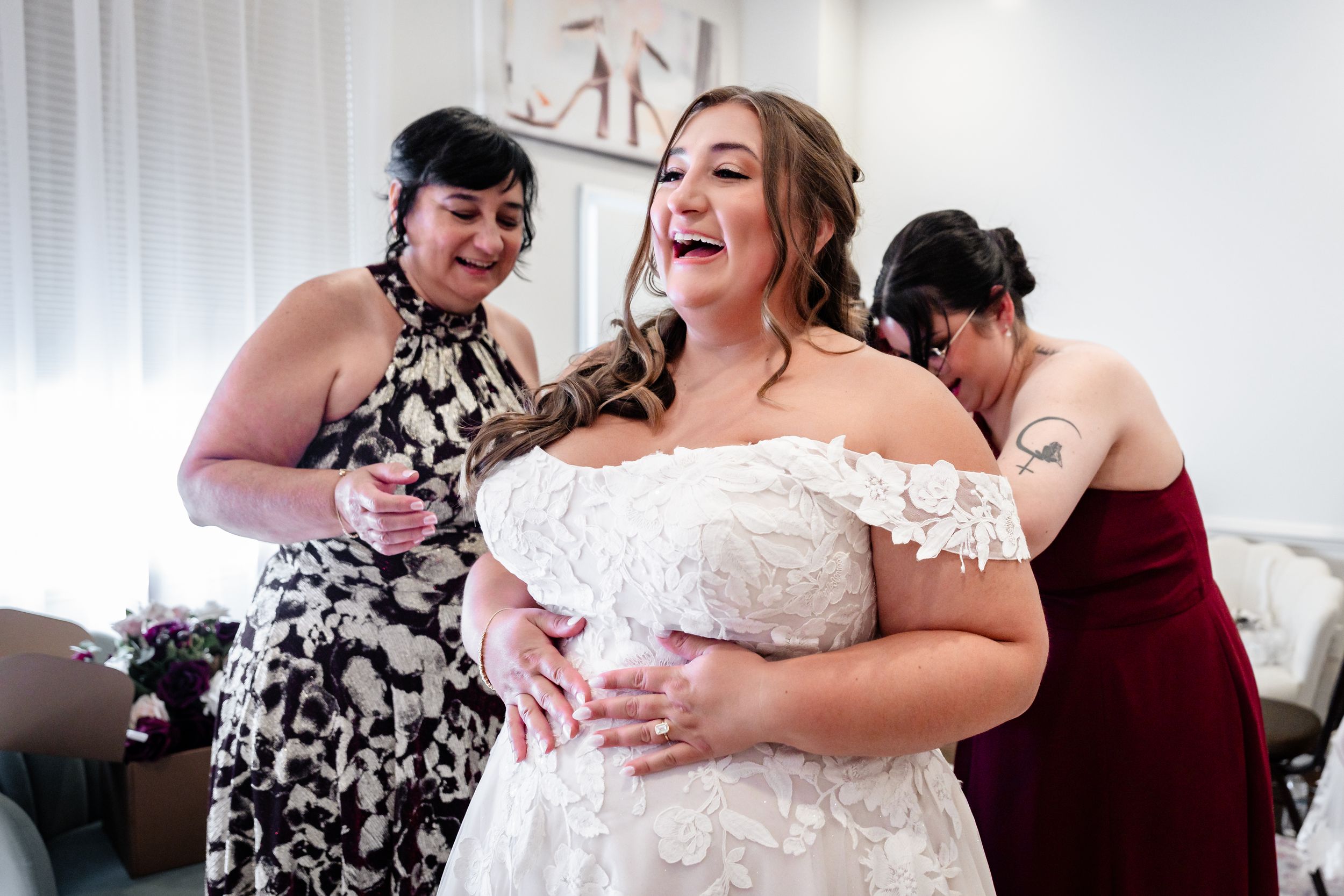 Bride laughing joyfully as bridesmaids help her into a white lace off-shoulder wedding dress before the ceremony.