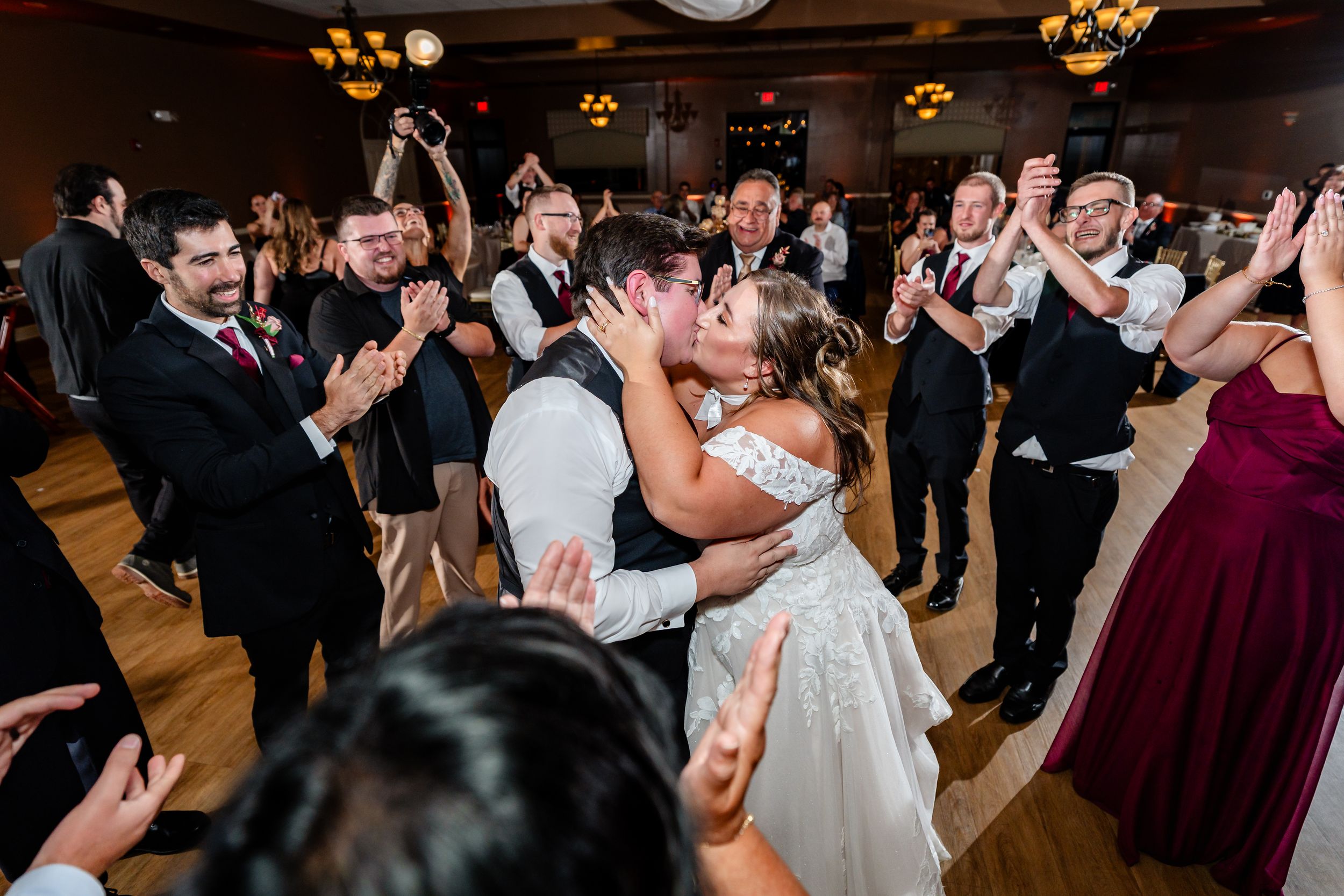 Bride and groom share a passionate kiss surrounded by cheering guests on the dance floor.