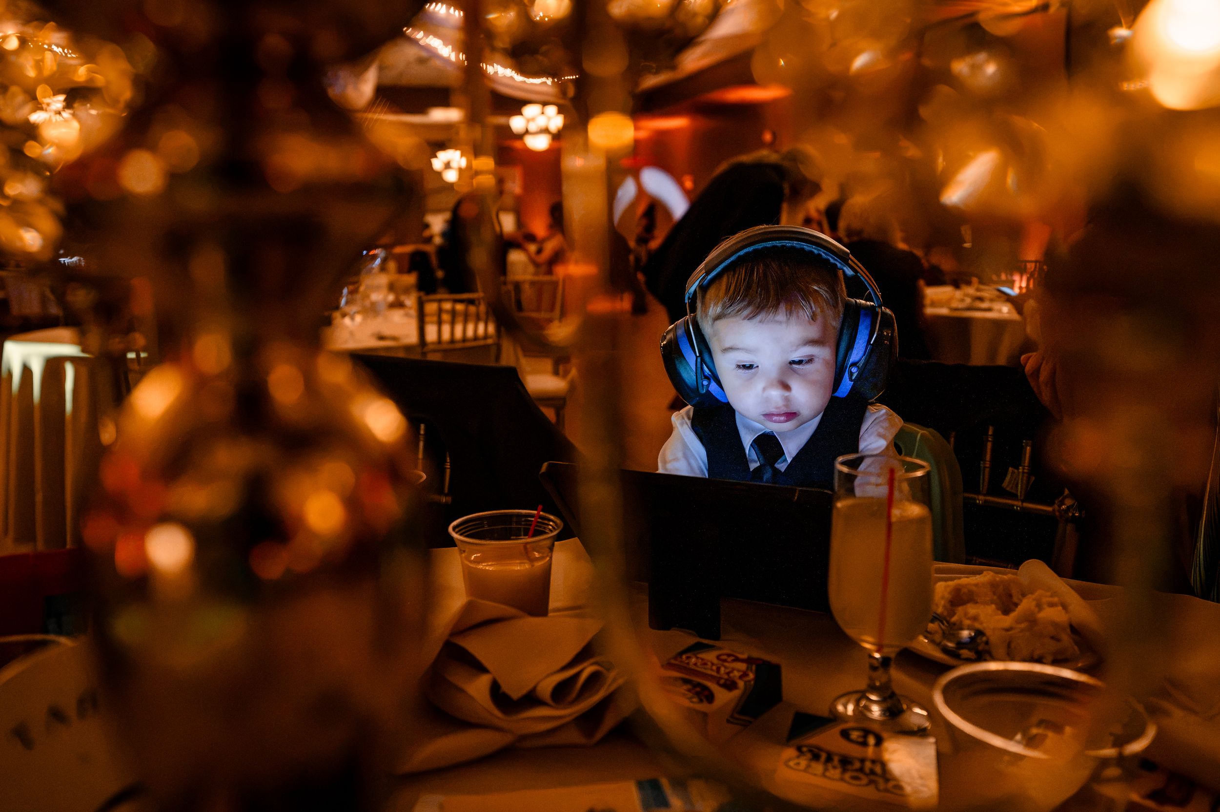 Young child wearing blue headphones watching a tablet at a dimly lit wedding reception table.
