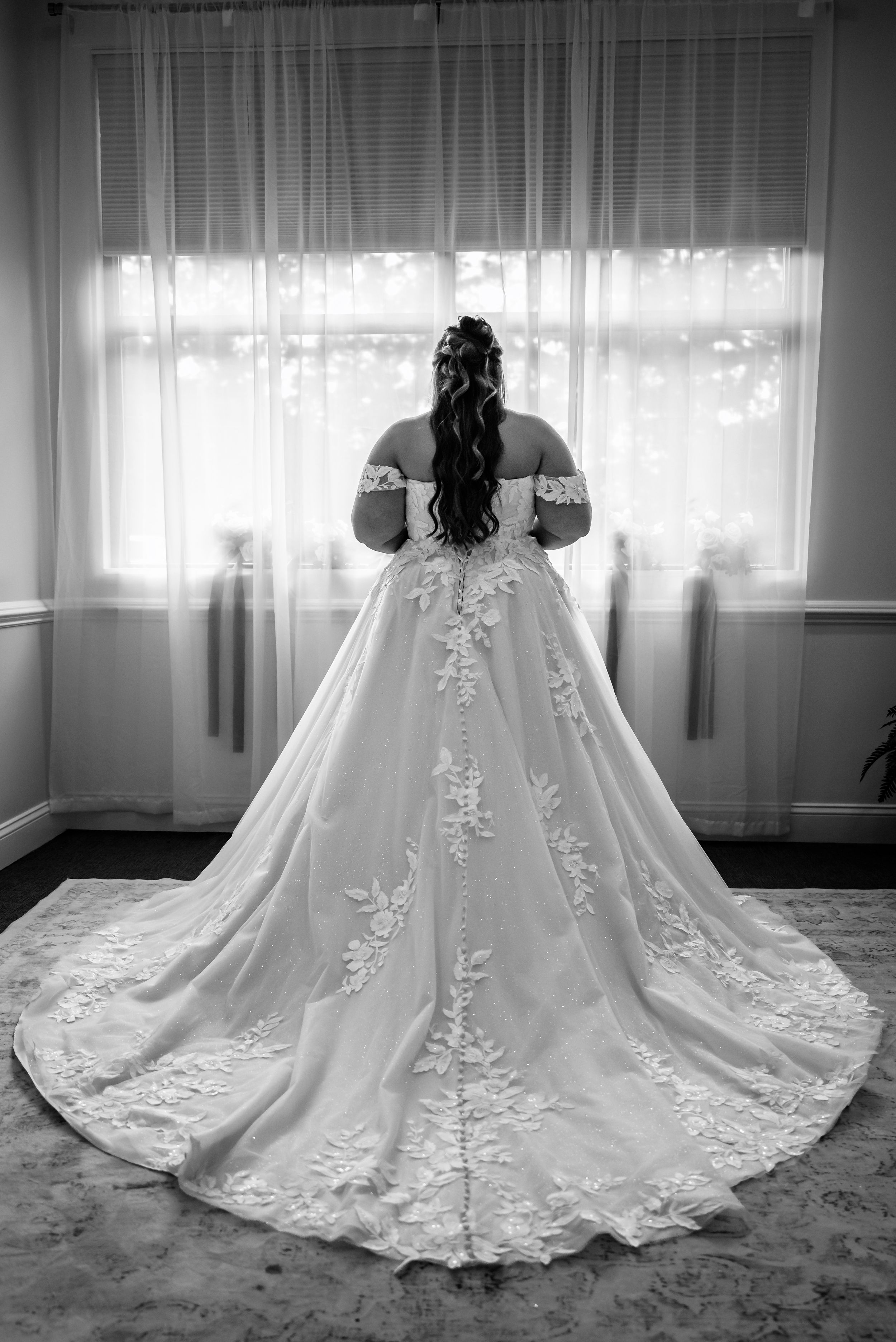 Bride in elegant off-shoulder ball gown with lace train standing before window in black and white.
