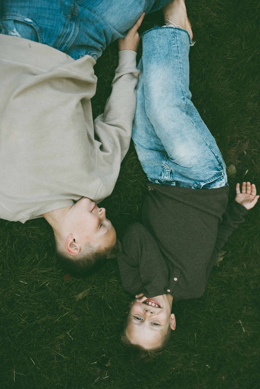 Two children lying on grass looking up, wearing jeans and casual tops in a candid outdoor portrait.