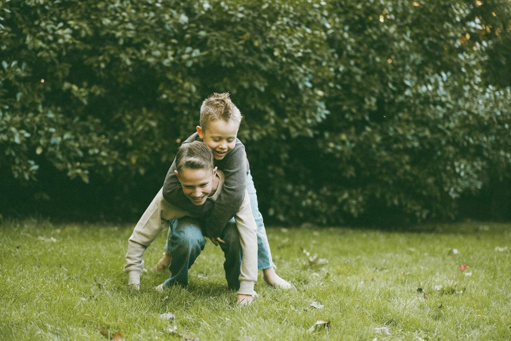A mother and young child playing piggyback on green grass in front of lush garden hedges outdoors.