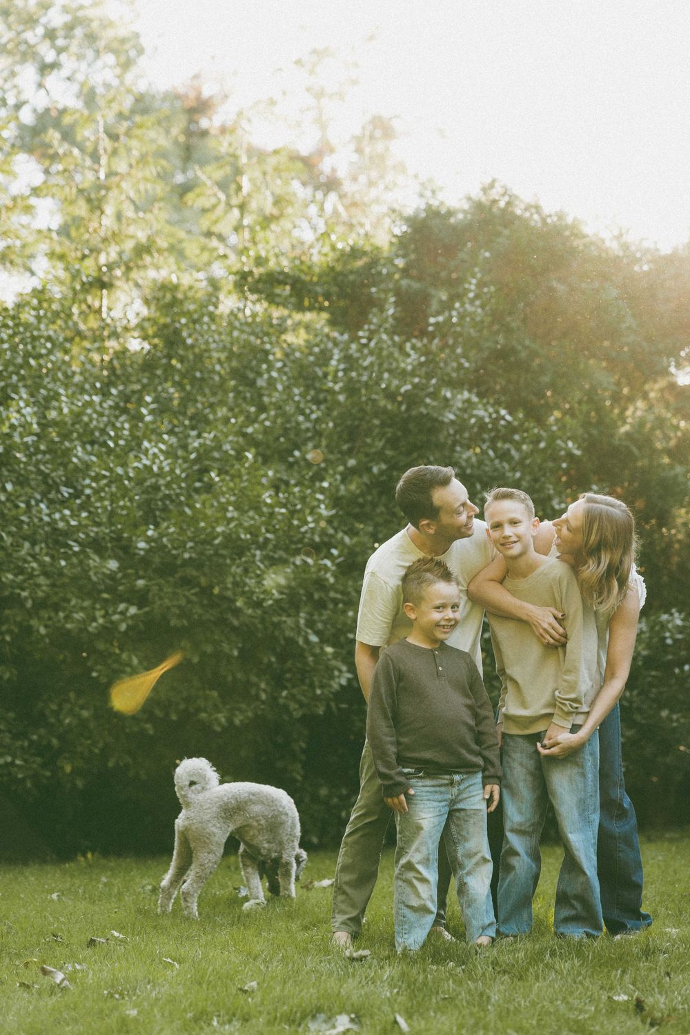 Family of four posing together outdoors on green grass with a white poodle dog beside them near lush trees.
