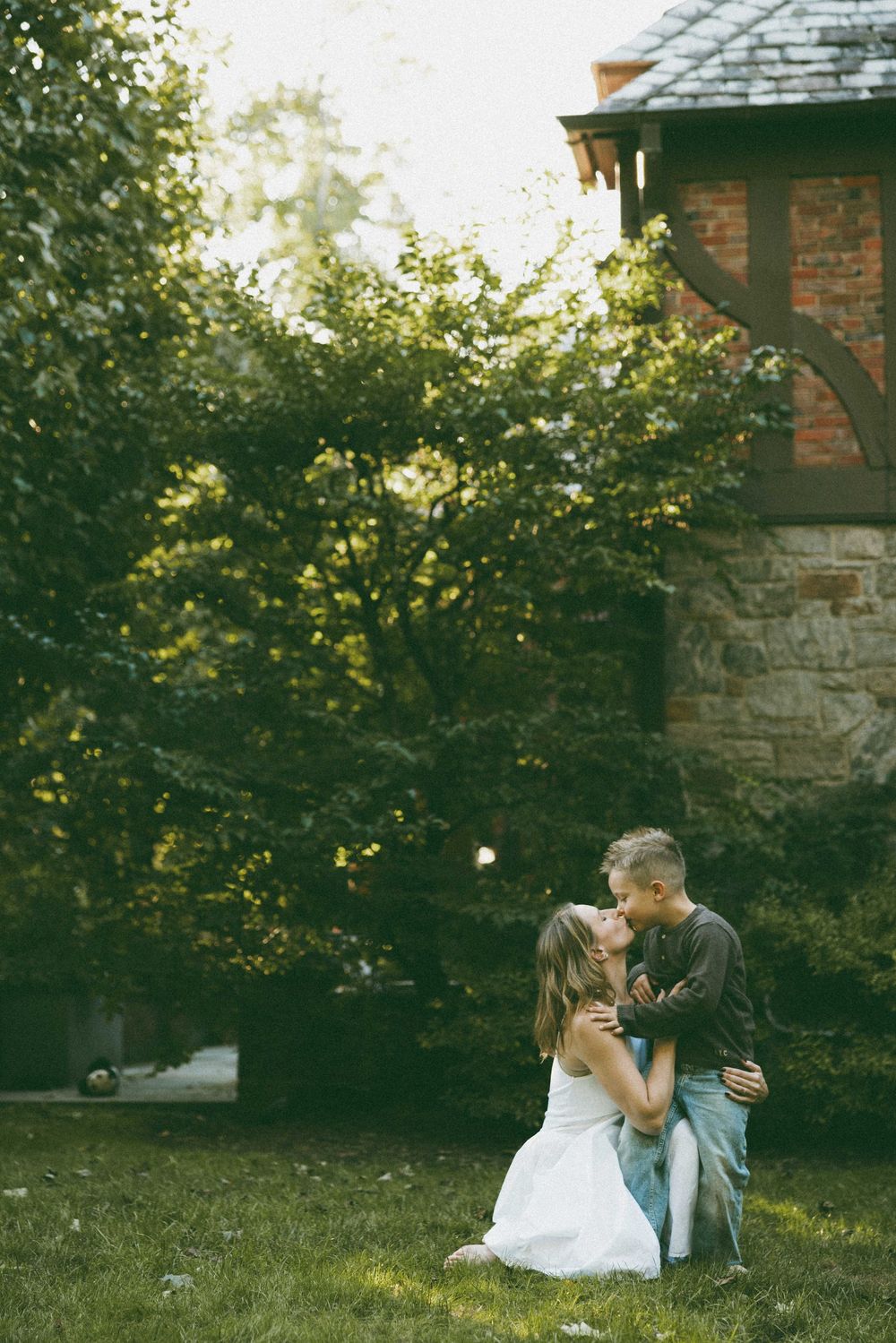 A couple shares a tender embrace on green grass beside a stone building surrounded by lush trees.
