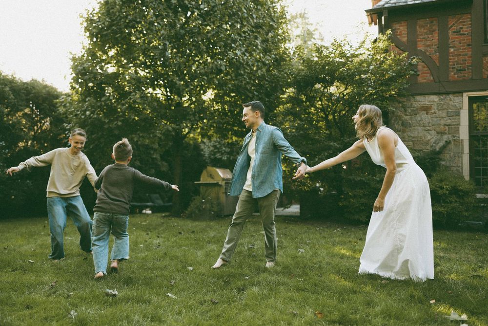 A joyful family plays together in a lush green backyard, with a bride in a white dress holding hands with children.