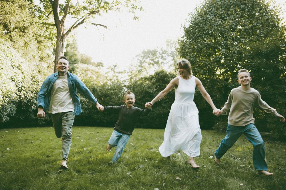 A joyful family of four holds hands and jumps together in a lush green garden on a sunny day.