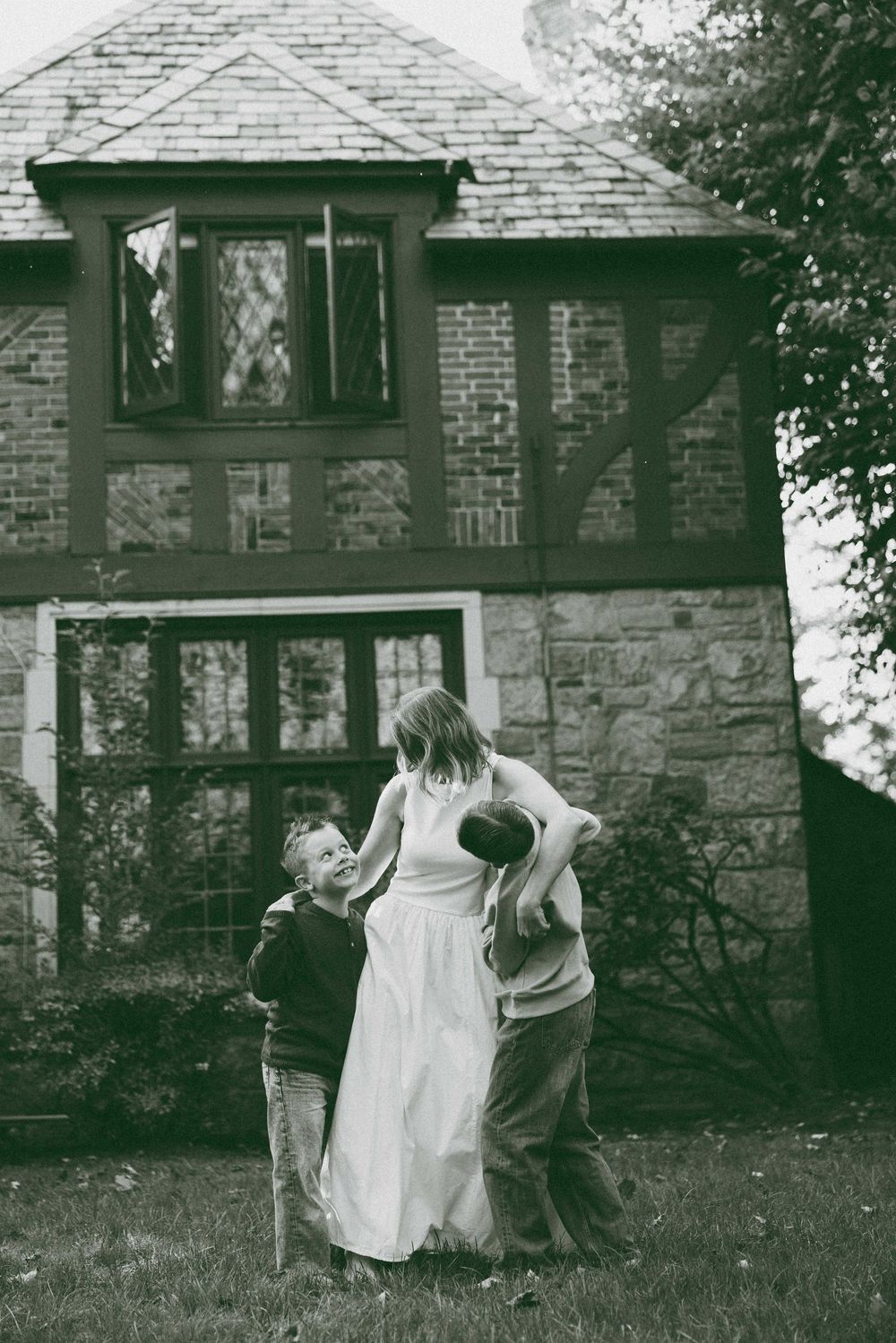 Black and white photo of a woman in a white dress with two children playing in a yard in front of a brick Tudor-style home.
