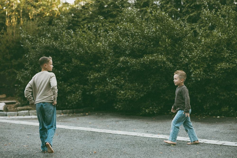 Two young boys walking on a road with lush green trees in the background.