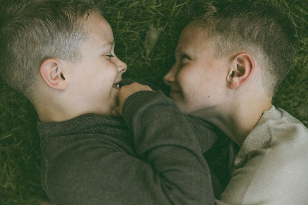Two young boys with short hair lying on grass, laughing and facing each other in a candid outdoor moment.