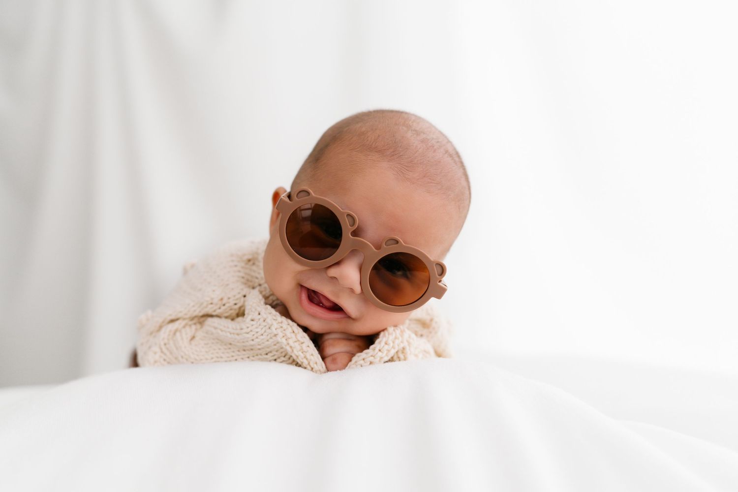 Adorable newborn baby wearing oversized brown round sunglasses, lying on a white blanket in a bright studio setting.