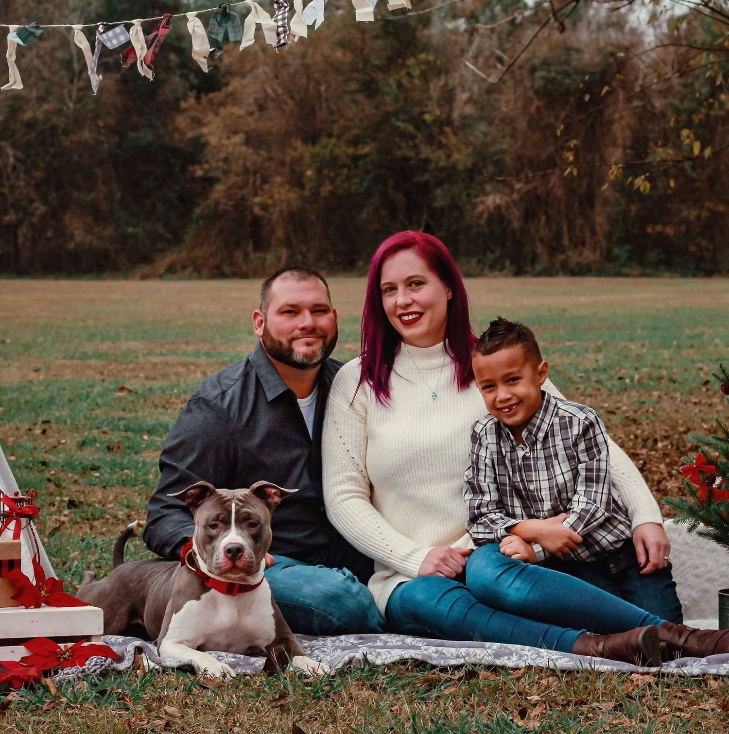Family of three poses with gray pit bull dog on blanket in park with holiday decorations for Christmas photo.