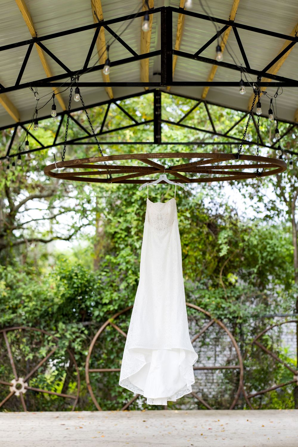 White wedding dress hanging under a rustic outdoor pavilion with string lights and lush green garden backdrop.