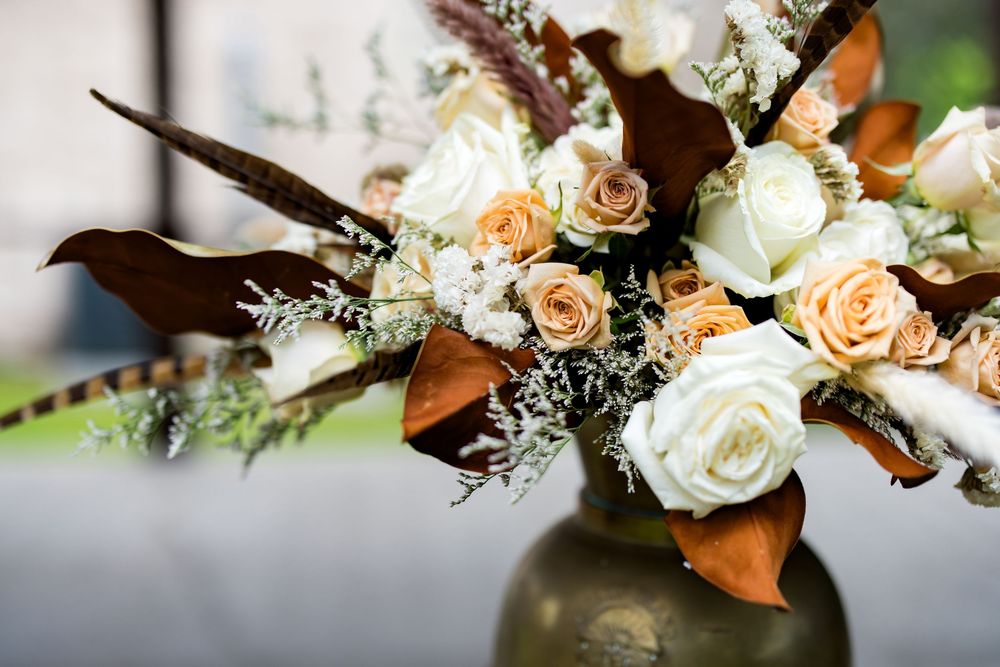 Elegant bridal bouquet with white and peach roses, brown calla lilies, and feathers in a gold vase.