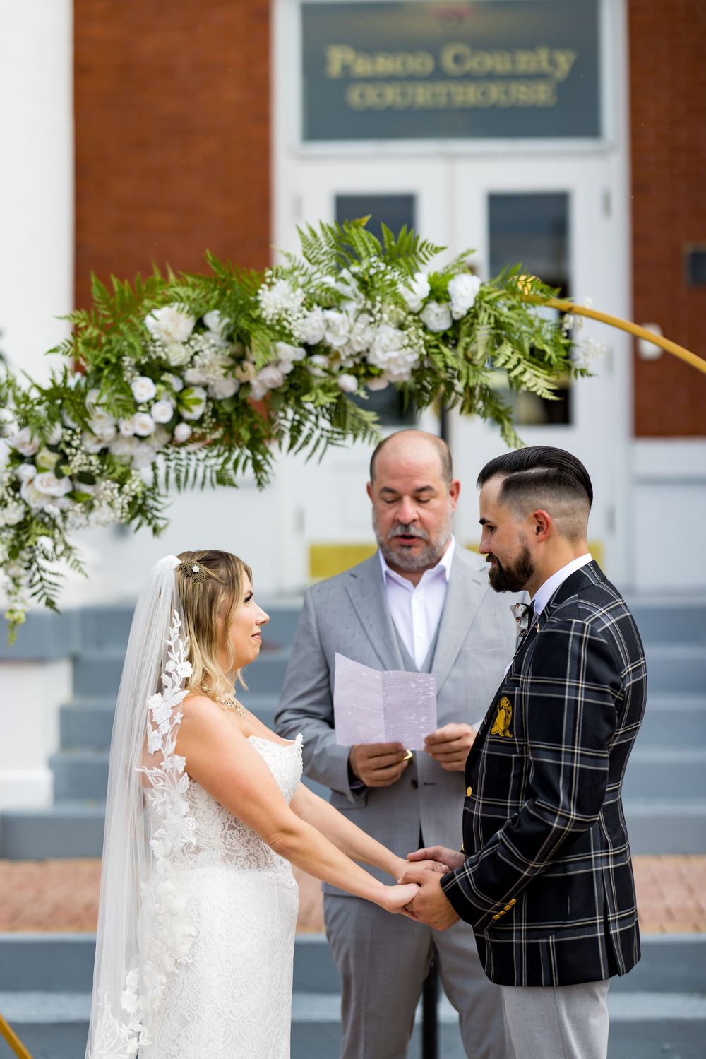 Couple exchanges vows at Pasco County Courthouse wedding ceremony with floral arch and officiant reading.