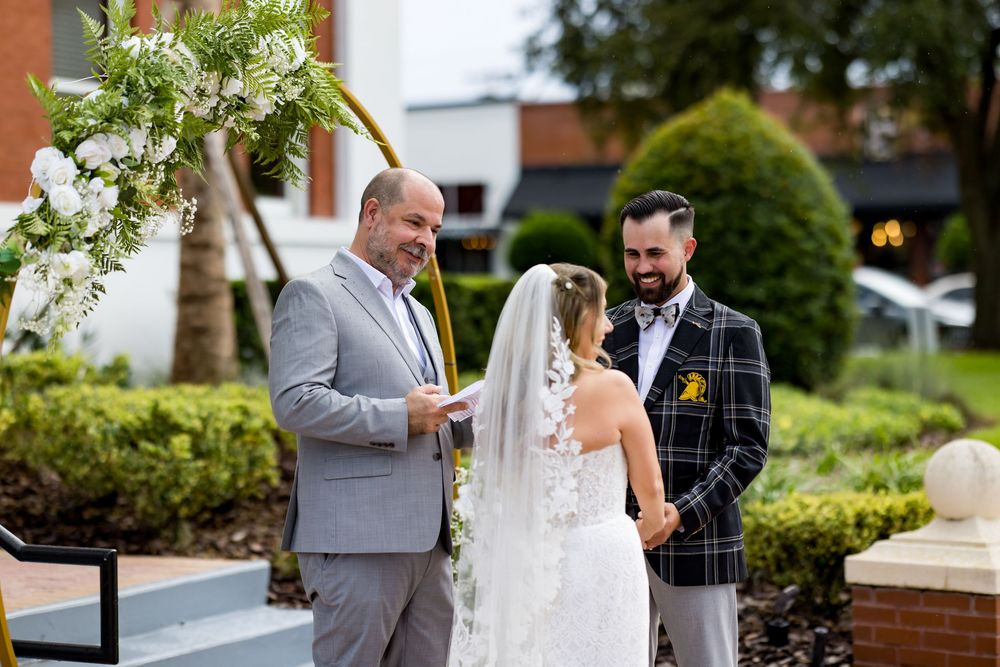 Outdoor wedding ceremony with officiant reading vows to bride in lace veil and groom in plaid suit, surrounded by floral arch.