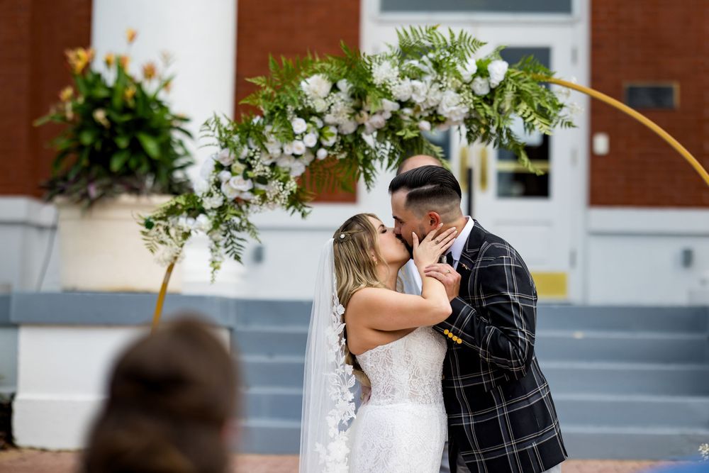 Bride and groom share first kiss under floral arch at outdoor wedding ceremony.