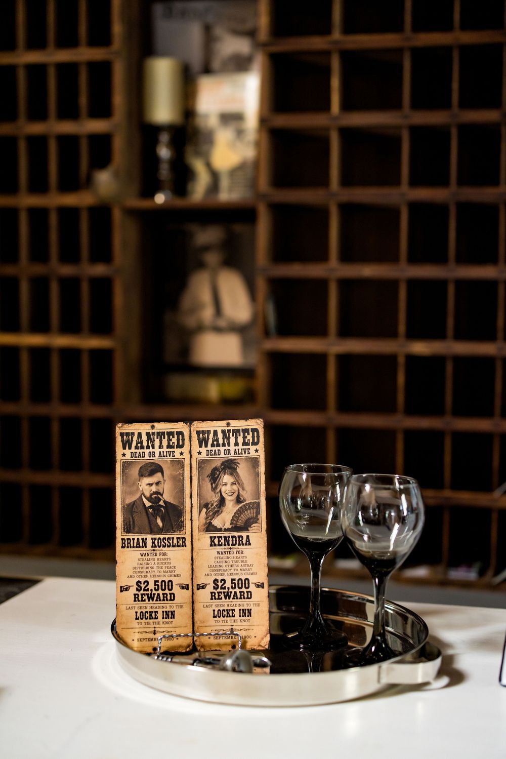 Western-themed wanted posters for Brian Kossler and Kendra with $2,500 reward at Locks Inn beside wine glasses.