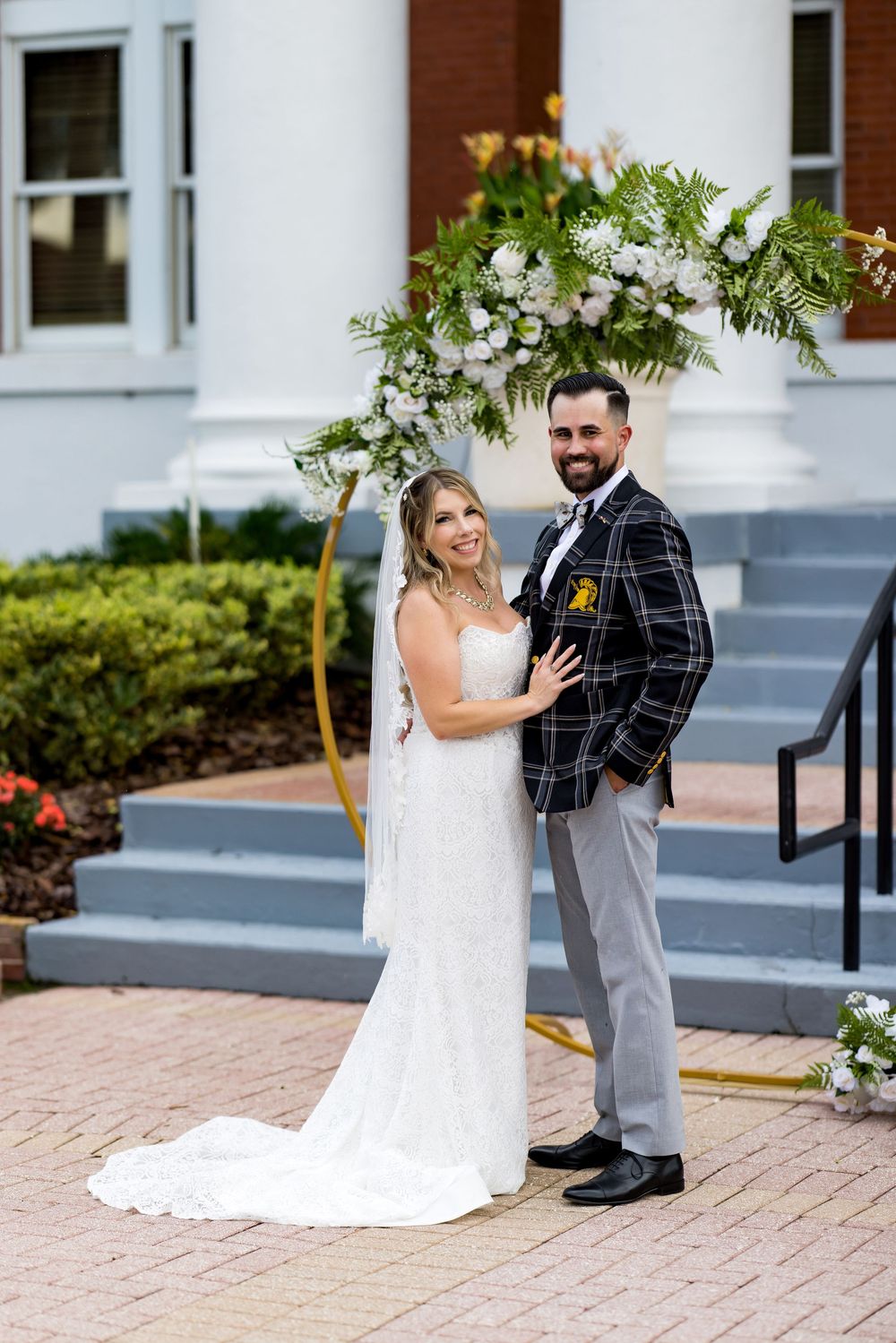 Bride in white gown and groom in plaid blazer pose under floral arch on brick path outside white building.