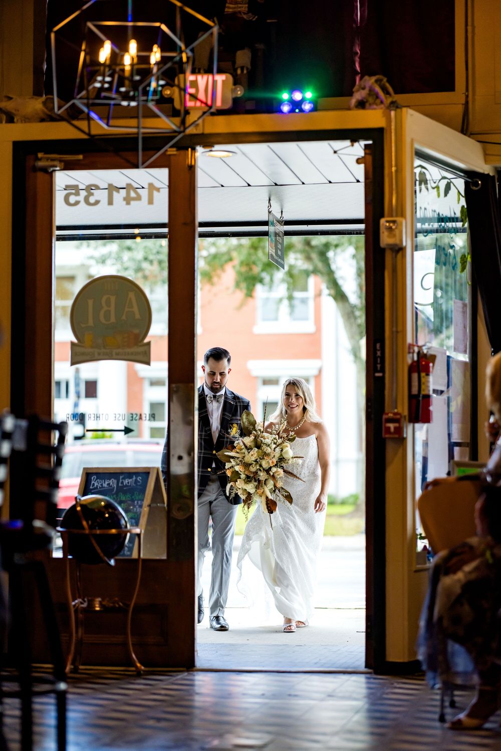 Bride and groom entering a venue through open doors, bride holding a large bouquet, warm indoor lighting.