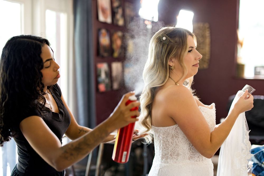A stylist sprays hairspray on a bride in a white wedding dress while another assistant helps her get ready.