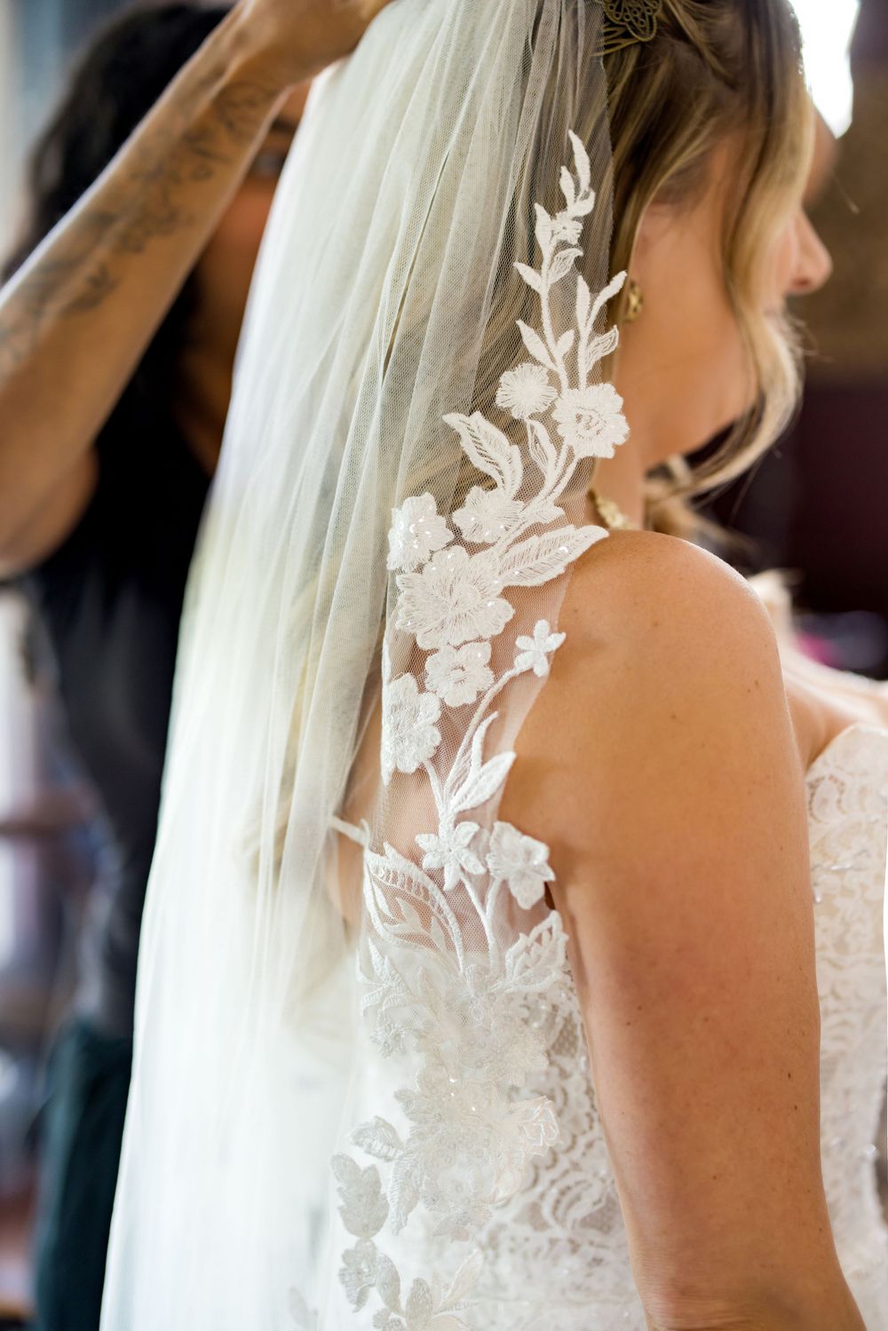 Bride in lace wedding dress with floral veil being adjusted, showing intricate back detail.