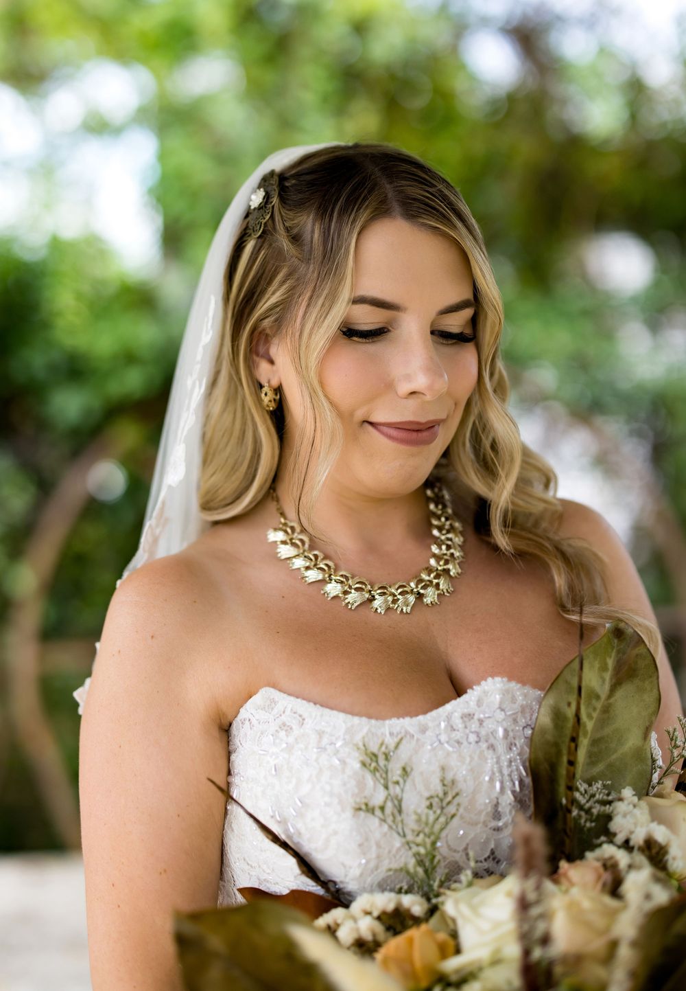 Bride in strapless lace gown with gold leaf necklace and veil holds bouquet, looking down gracefully outdoors.